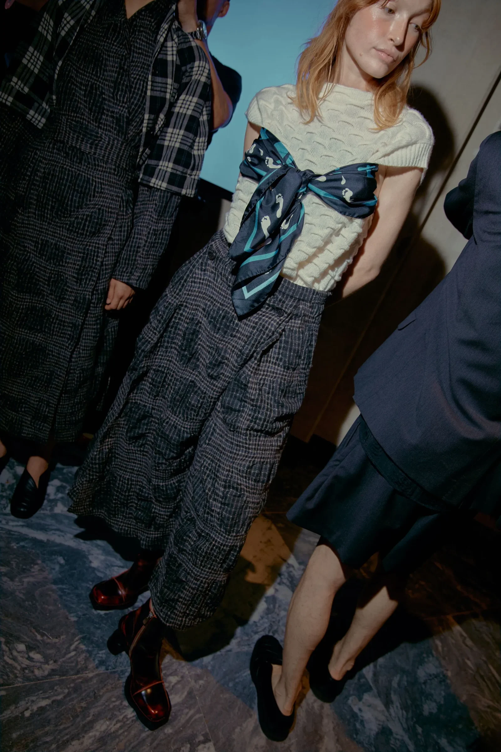 Models wearing plaid outfits with pleated skirts and a large blue scarf bow at a fashion-show backdrop.
