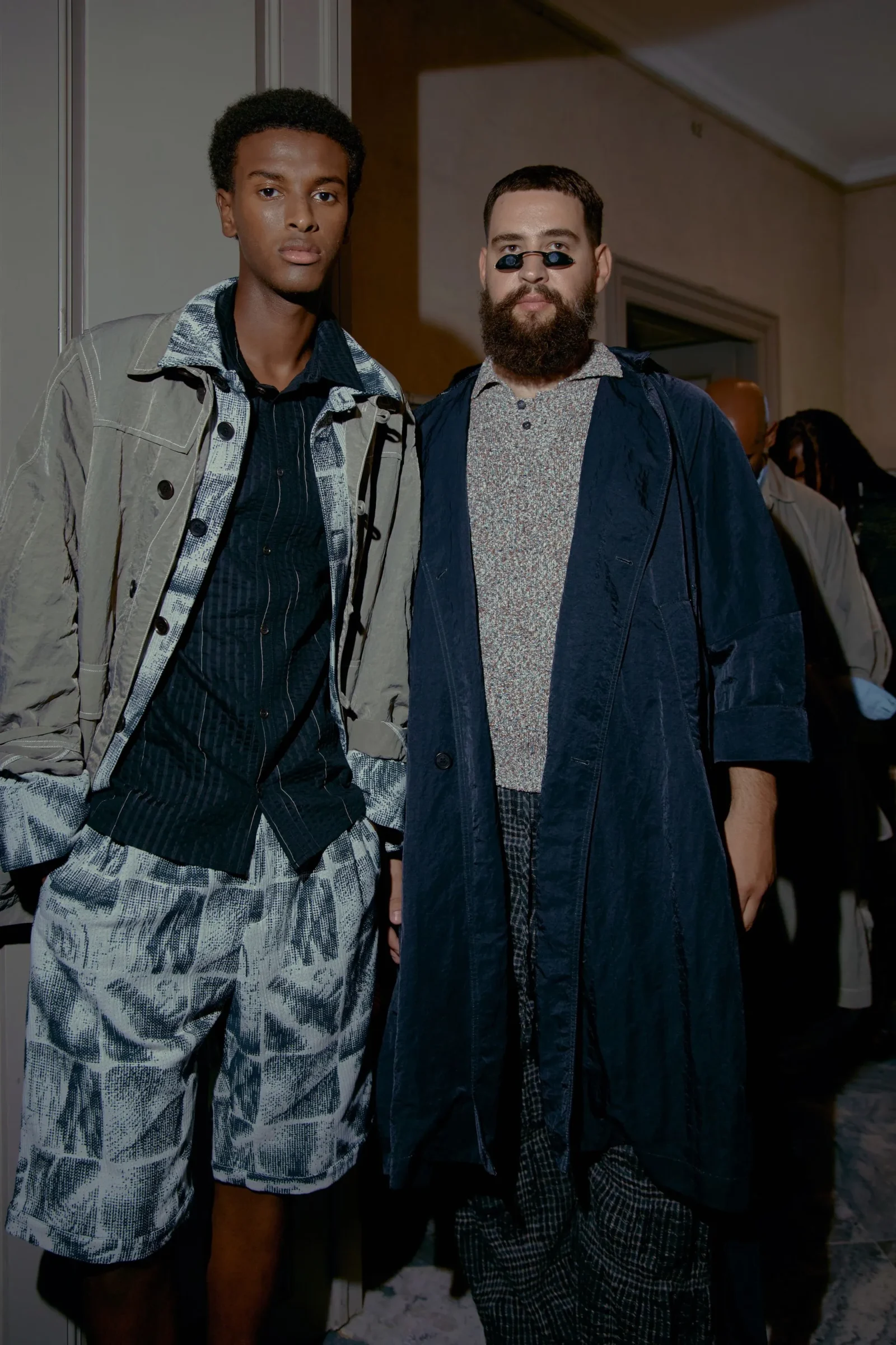 Two men pose indoors in stylish layered outfits: one in patterned shorts and a light jacket, the other with a beard and small sunglasses under a dark coat.