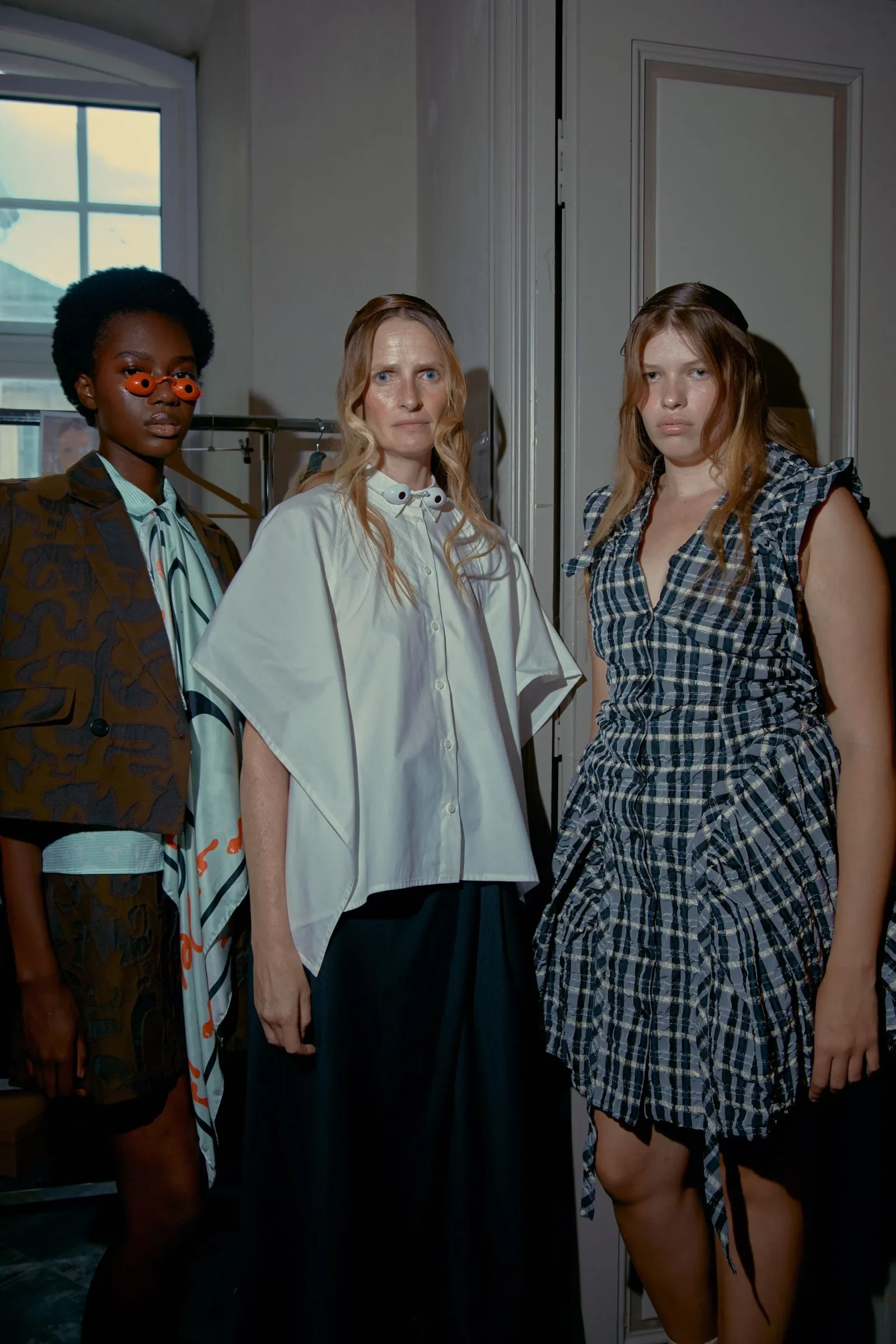 Three fashion models pose indoors in eclectic outfits: left wears patterned blazer with orange sunglasses, center in oversized white blouse, right in plaid dress.
