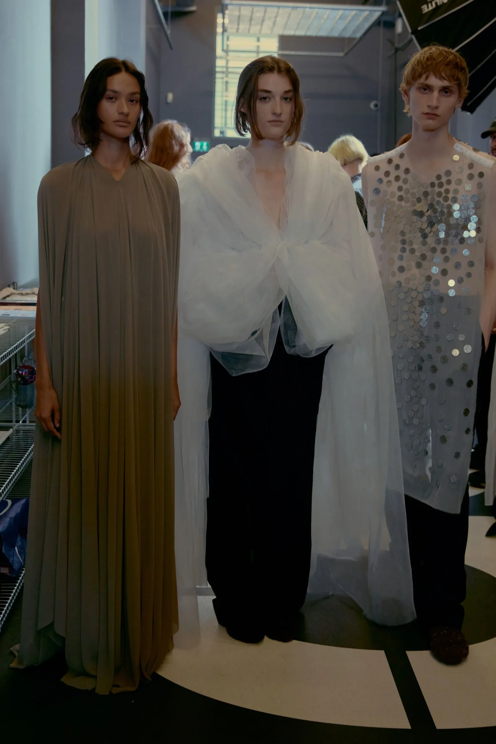 Three models pose in a fashion studio wearing dramatic gowns: left in a taupe pleated dress, center in a white voluminous blouse, right in a silver sequin sleeveless dress.