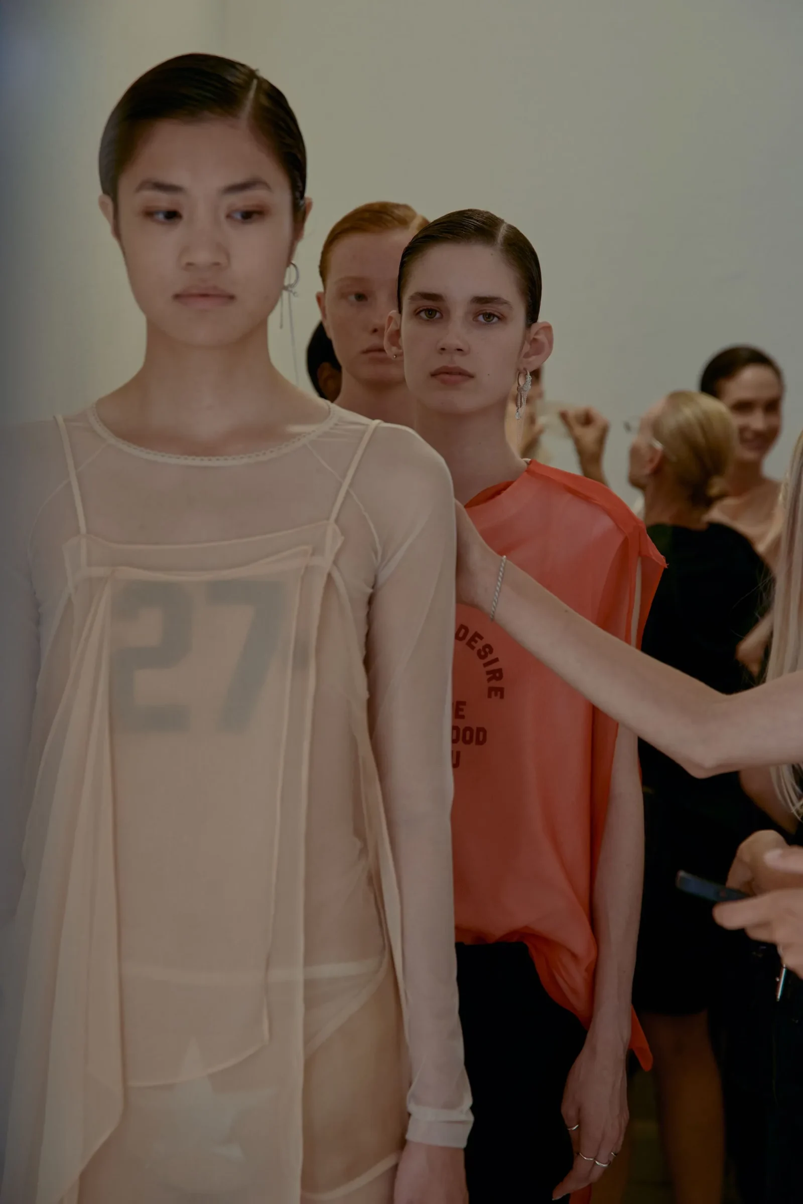 Models lined up backstage at a fashion show, wearing sheer beige and coral outfits