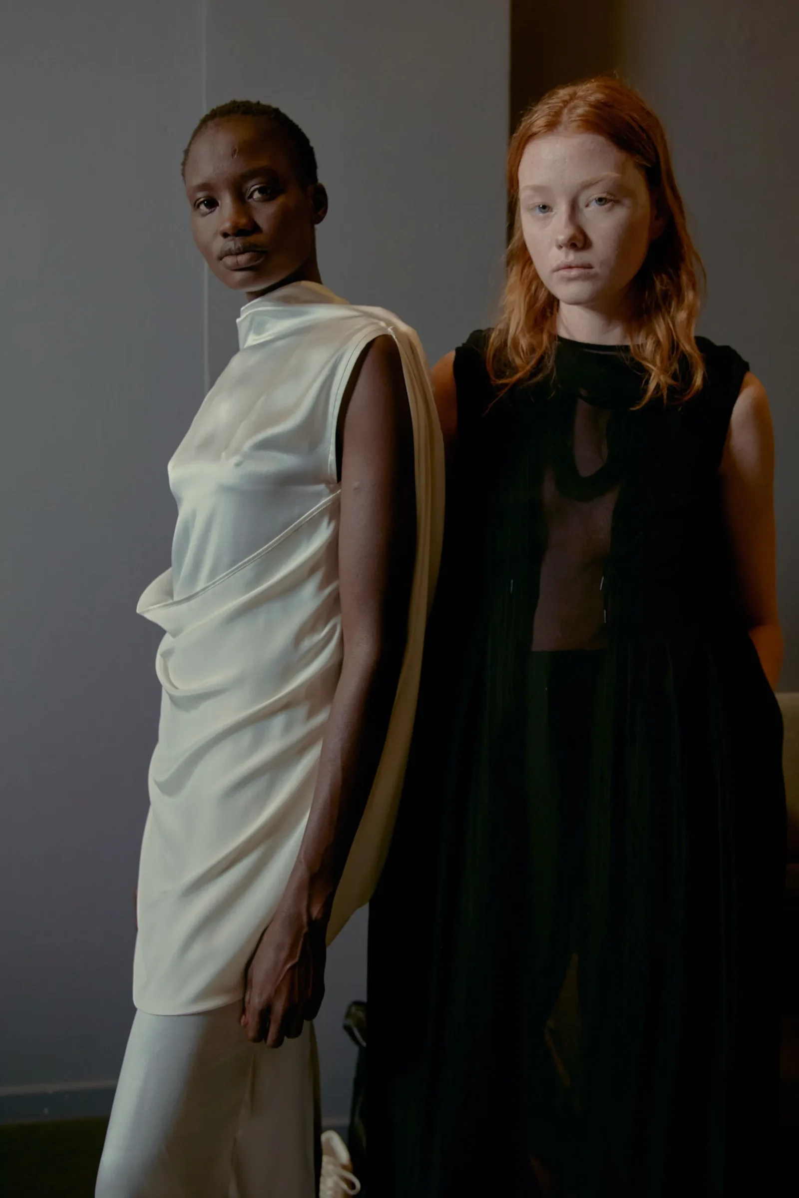 Two models stand side by side in a dim studio, one in a white draped satin dress on the left and one in a sheer black gown on the right, both facing the camera.