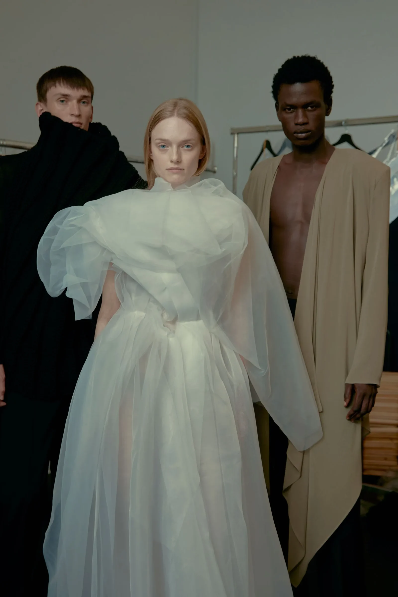 Three models pose in a studio wearing dramatic outfits: a central woman in a voluminous white gown, flanked by a man in black on the left and a man in beige on the right.