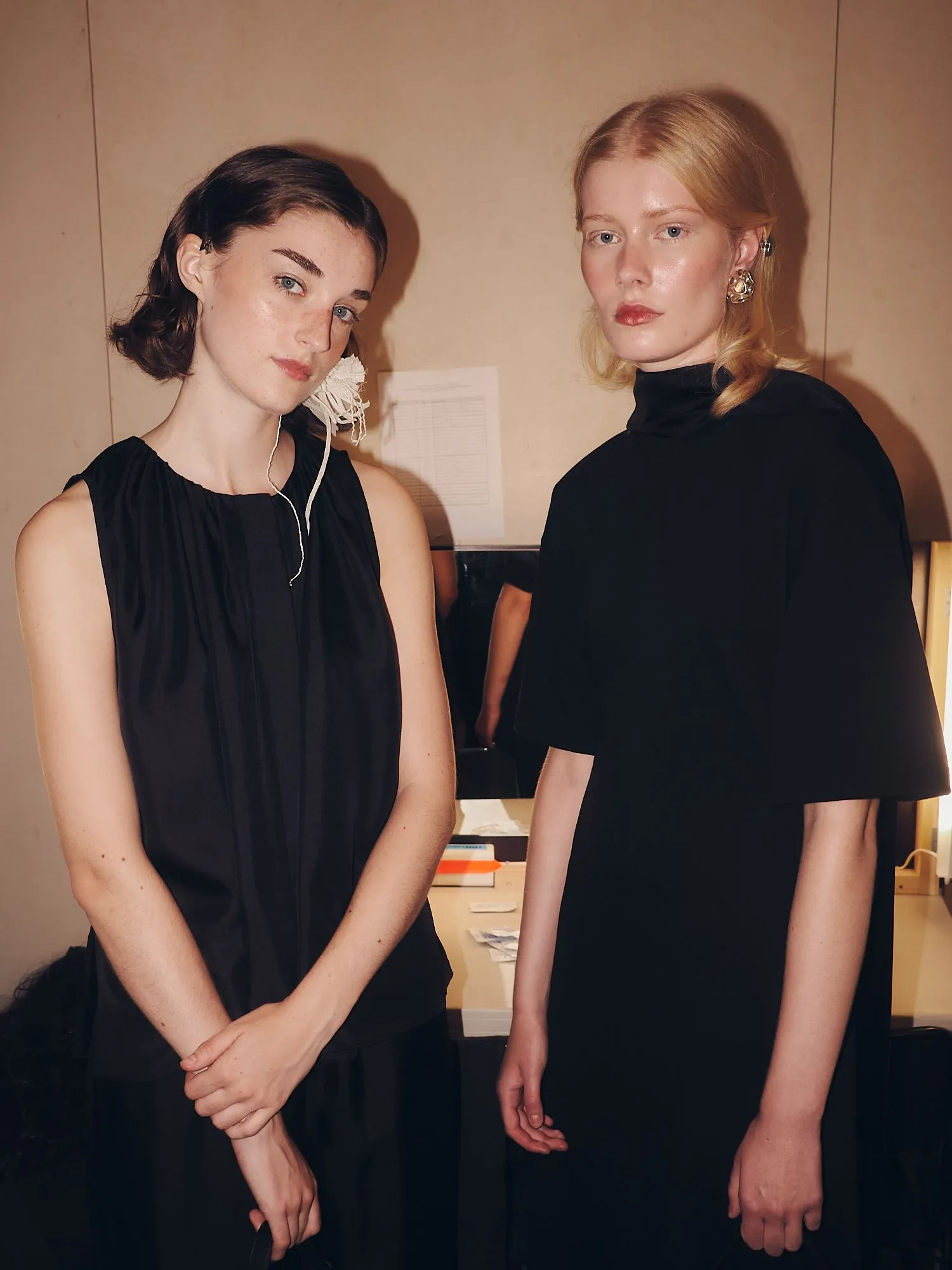 Two women in black outfits pose side by side indoors, one with short dark hair and the other with long blonde hair.