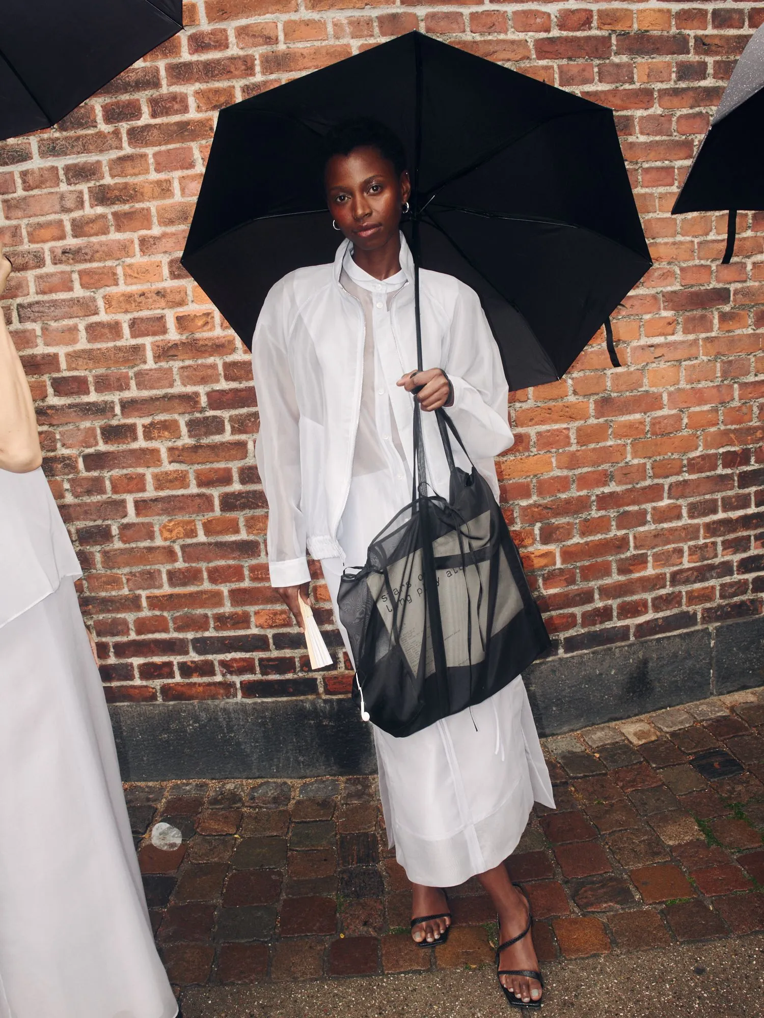 Person standing against a brick wall, holding a large black umbrella and wearing a white outfit with a sheer bag at the arm level.