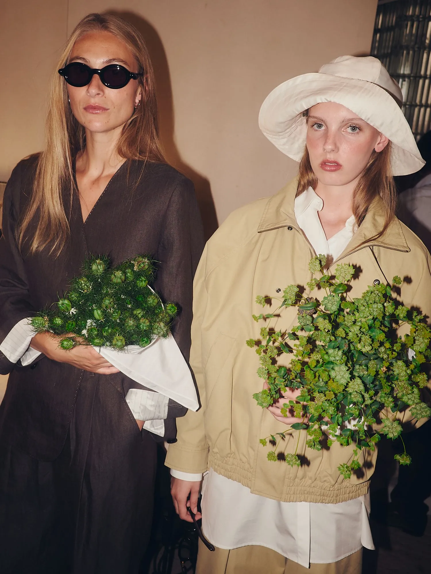 Two fashion models pose together; one in a dark dress with sunglasses, the other in a beige jacket and white hat, holding greenery.