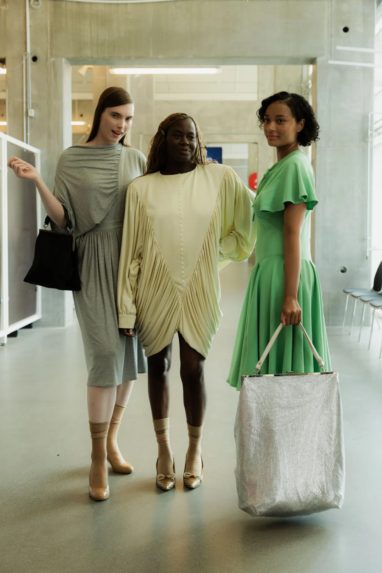 Three women in pastel dresses pose in a concrete-walled hallway, with a silver tote bag nearby.