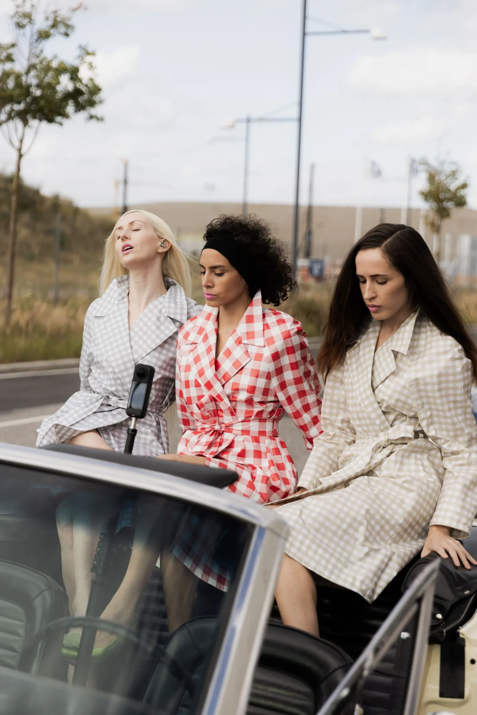 Three women in plaid coats pose beside a silver convertible in a parking lot.