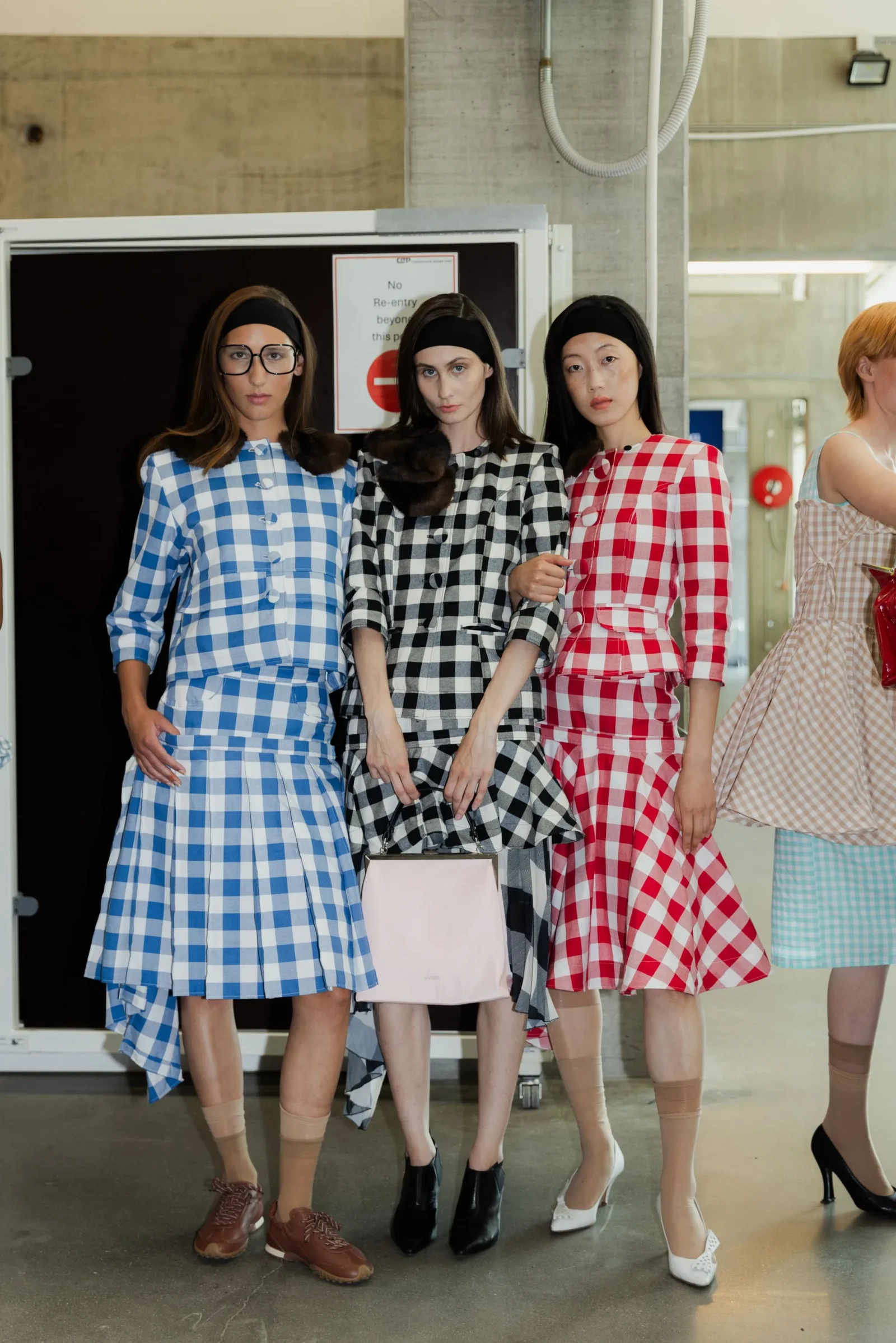 Three models stand in checkered gingham dresses (blue, black, red) posing on a runway backstage.