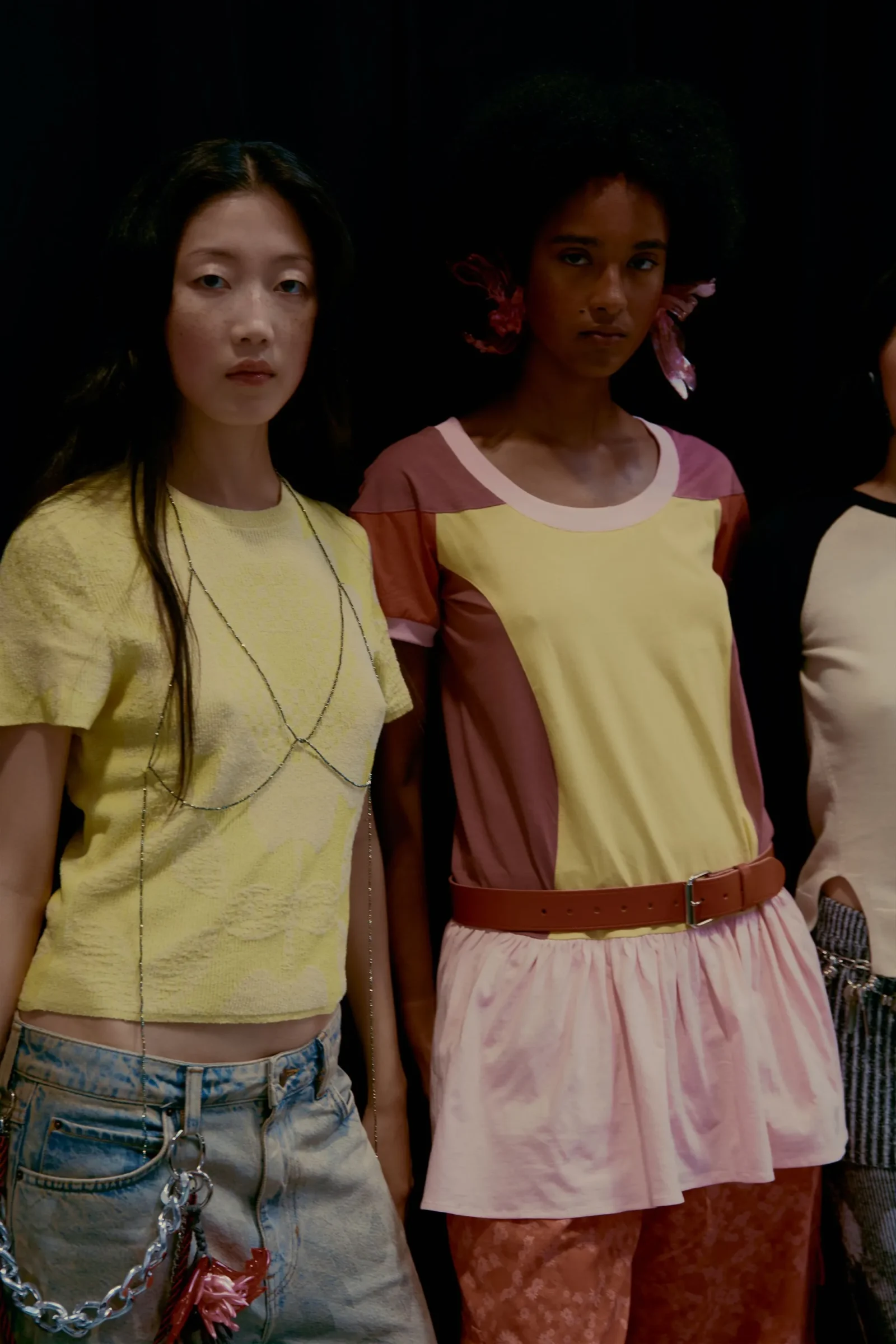 Two women pose in fashion outfits against a dark background, one in yellow top with layered chains and denim shorts, the other in a pastel color-block dress with a pink belt.