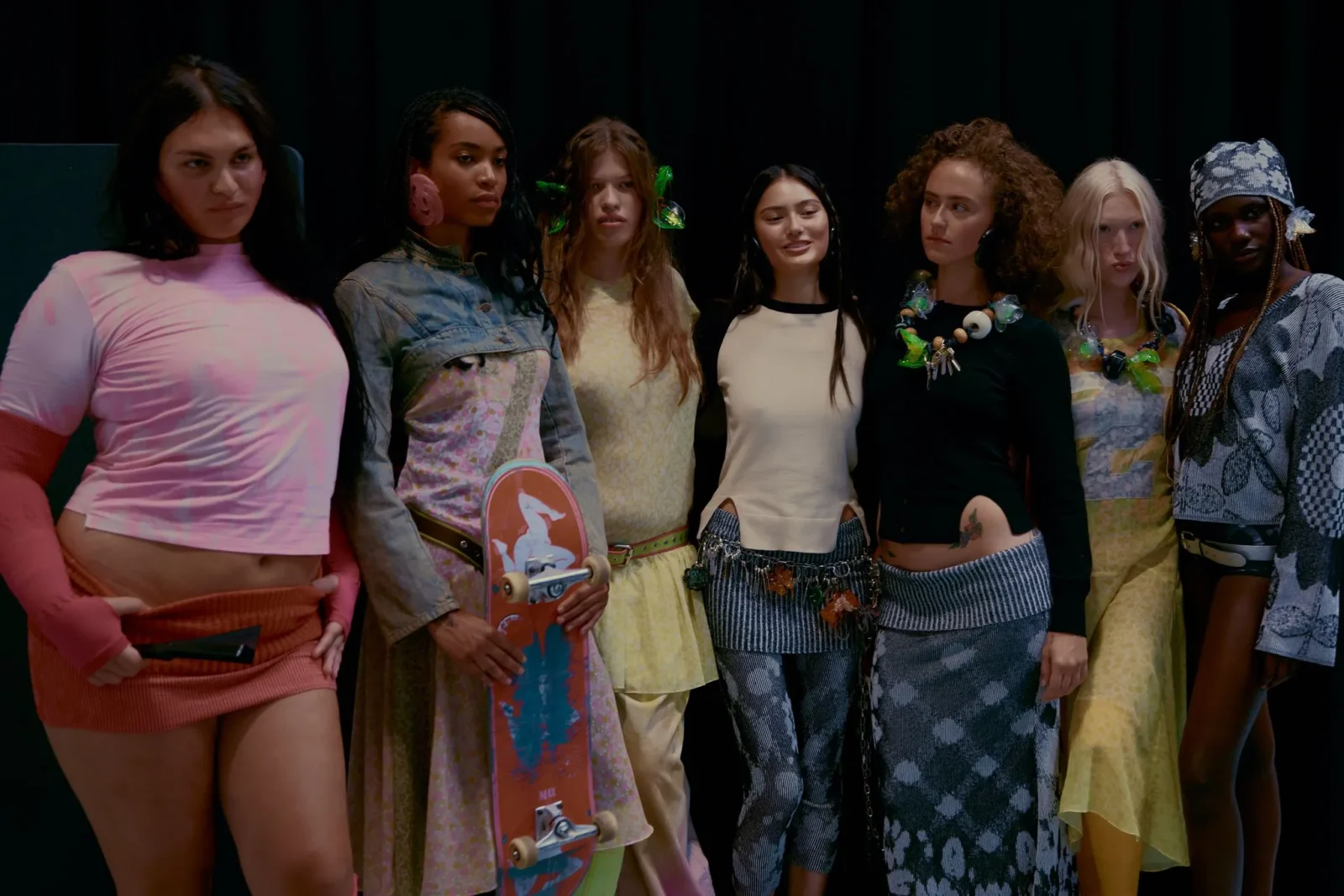 Eight young women pose together on a dark stage in colorful, trendy outfits and accessories, with a pink skateboard visible in the front left.