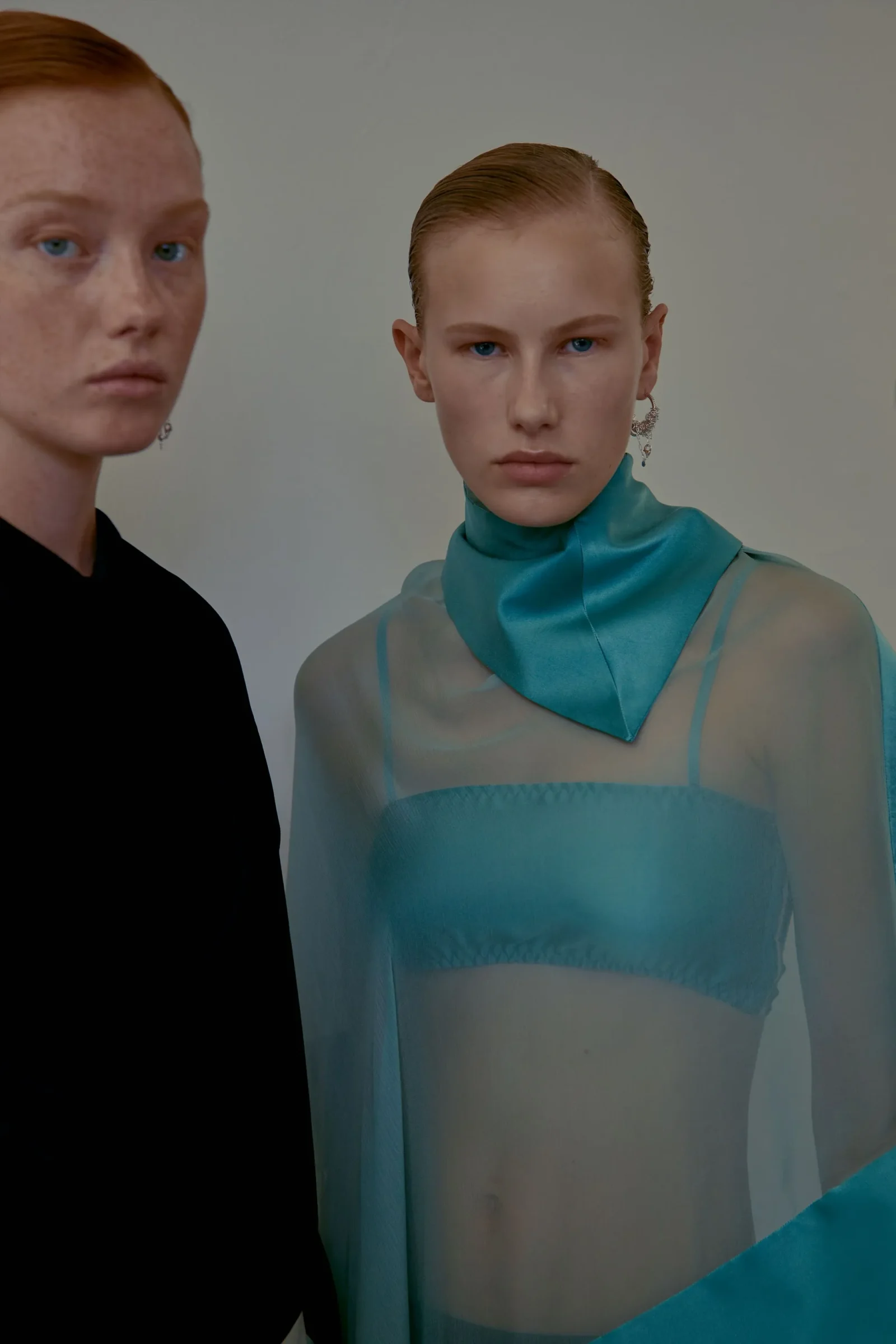 Two fashion models pose in a studio: a woman in a teal sheer top with slicked-back hair and dangling earrings stands beside another person in black.