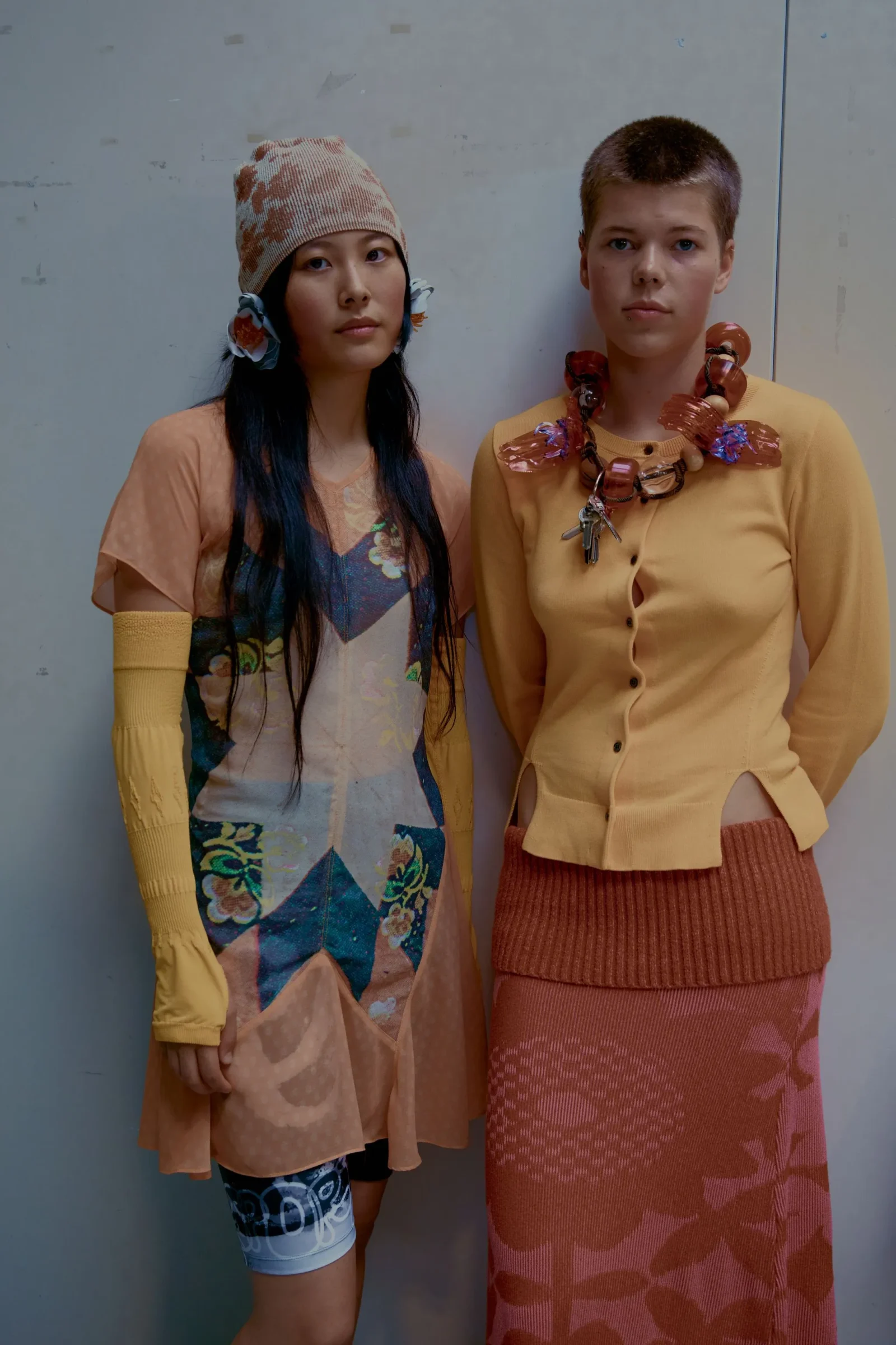 Two young people stand side by side against a pale wall, wearing colorful, patterned outfits and chunky accessories.