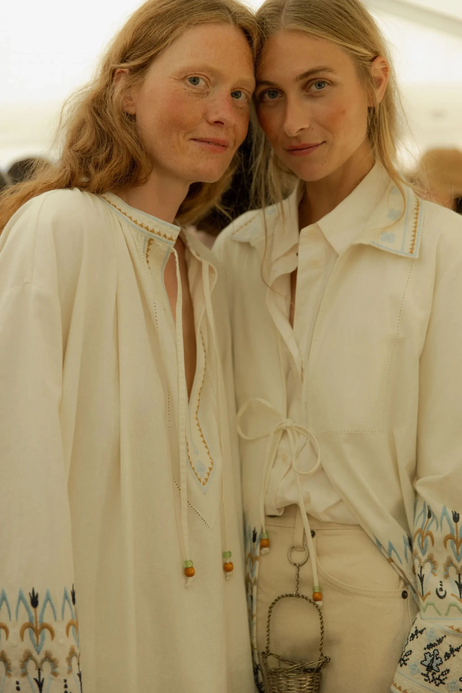 Two women in cream embroidered outfits pose closely together, smiling at the camera.