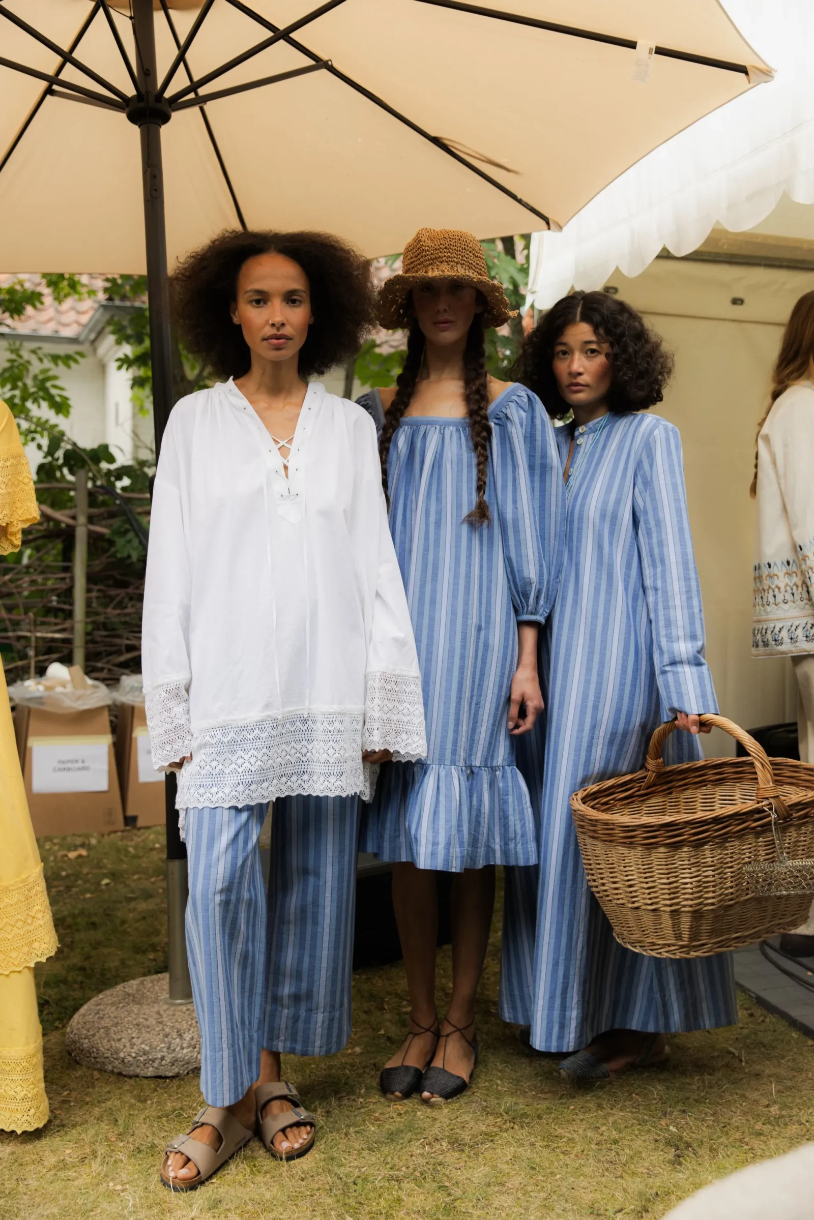 Three women stand under a large beige umbrella wearing blue and white striped dresses; one wears a white lace-trimmed blouse, and a wicker basket is visible nearby in a sunny outdoor setting.