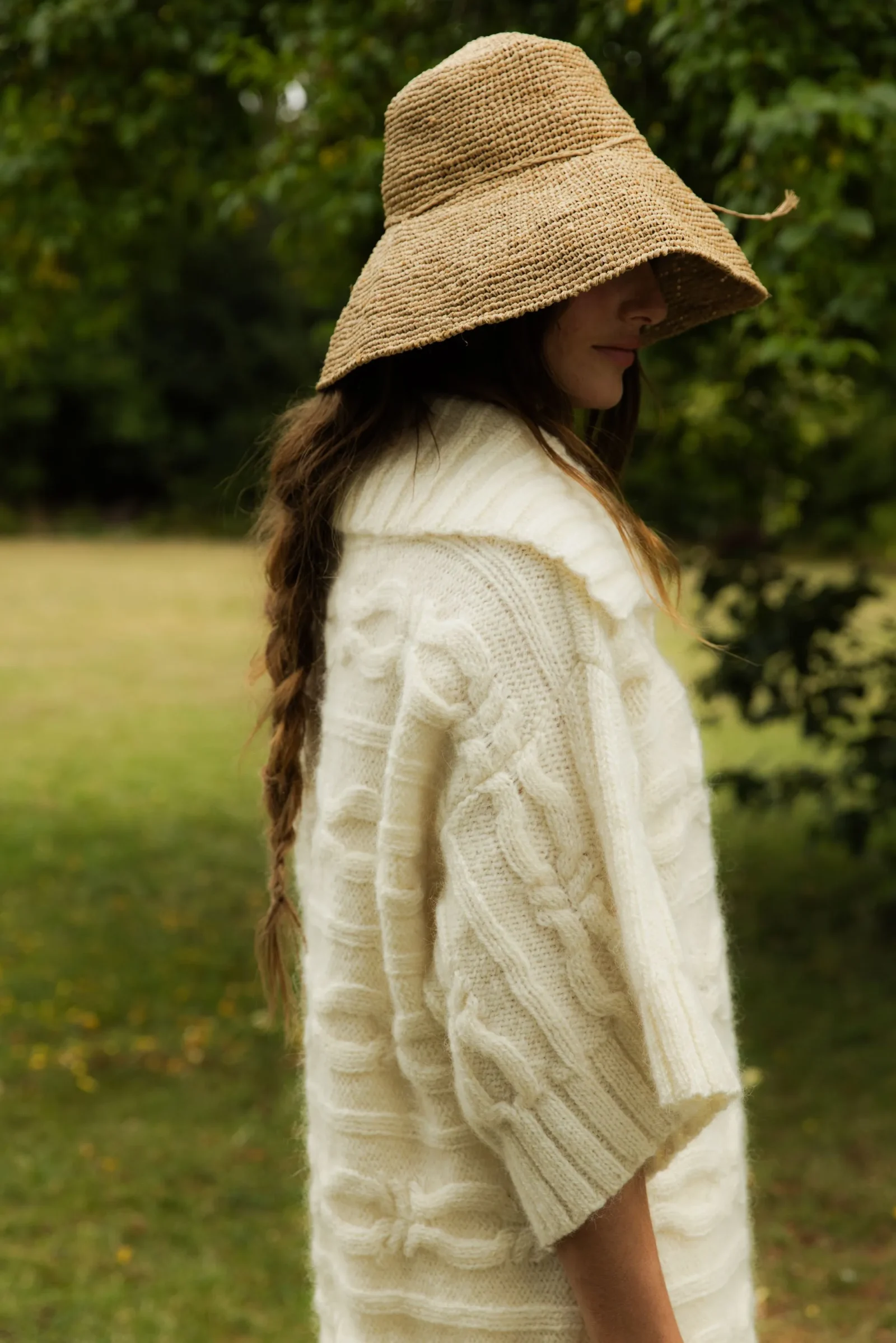Woman in a wide-brim straw hat and chunky cream sweater, seen in profile outdoors in a grassy, tree-filled area.