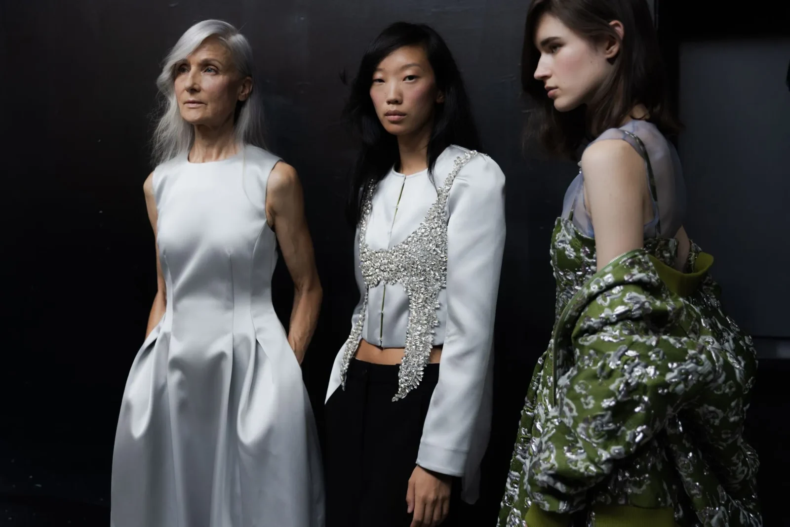 Three models pose against a dark backdrop in avant-garde outfits: white satin dress, silver-embellished vest, and green floral coat.