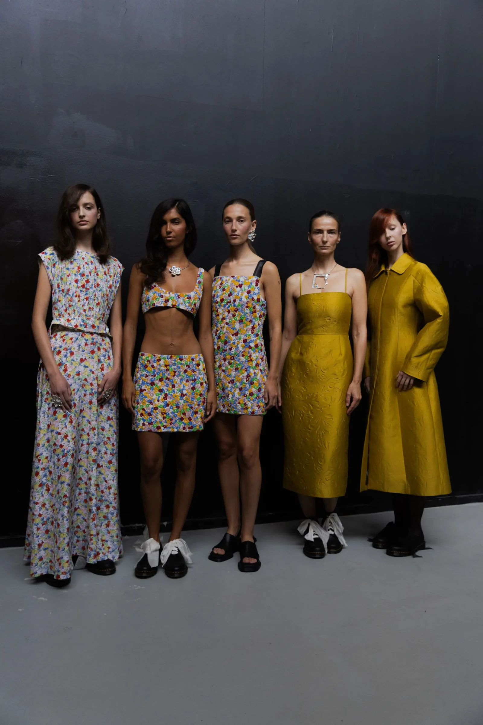 Five female models stand against a dark wall, wearing colorful floral outfits and a yellow dress, posing for a fashion shoot.