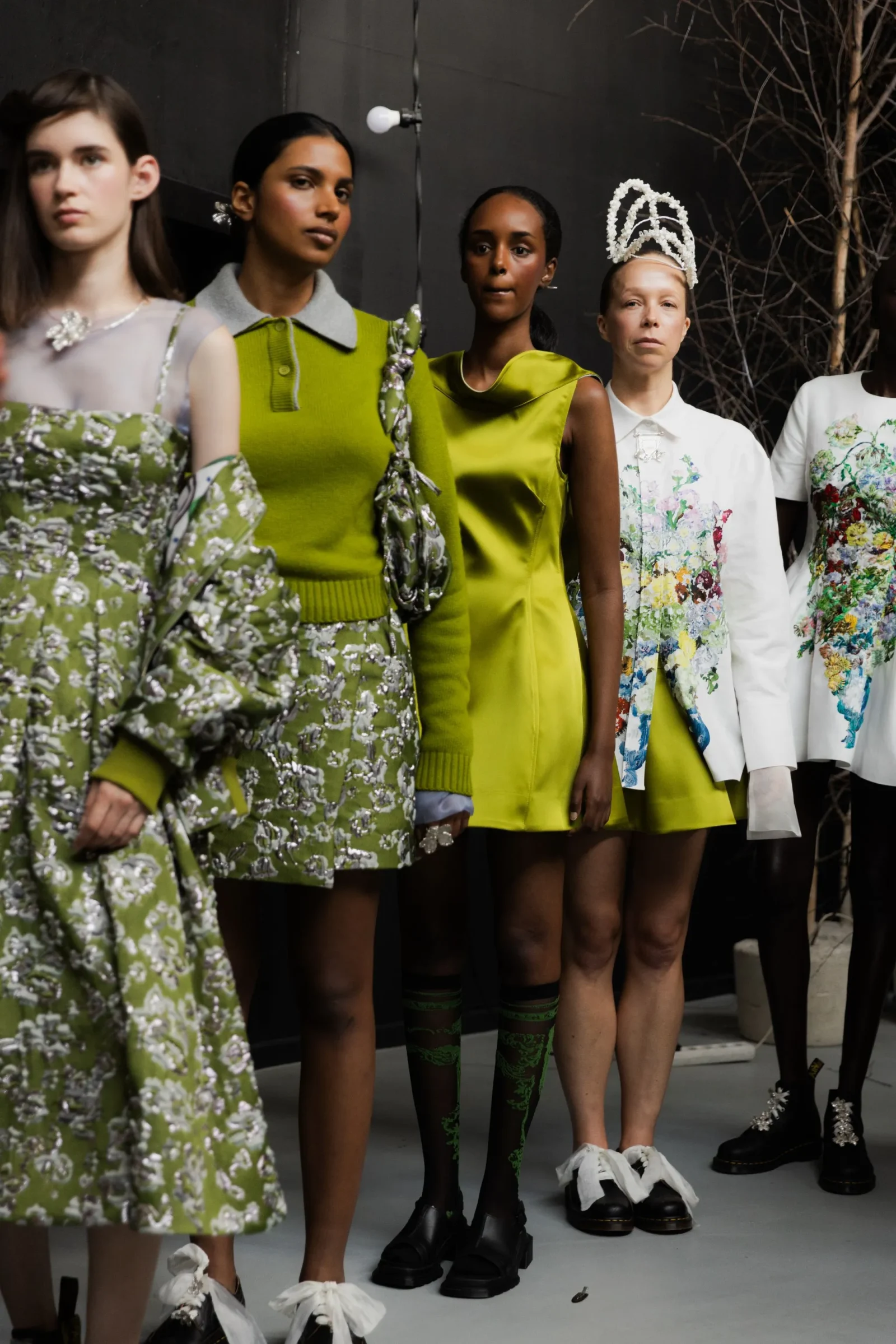 Line of models wearing bright green and floral outfits with decorative headpieces on a runway backdrop.