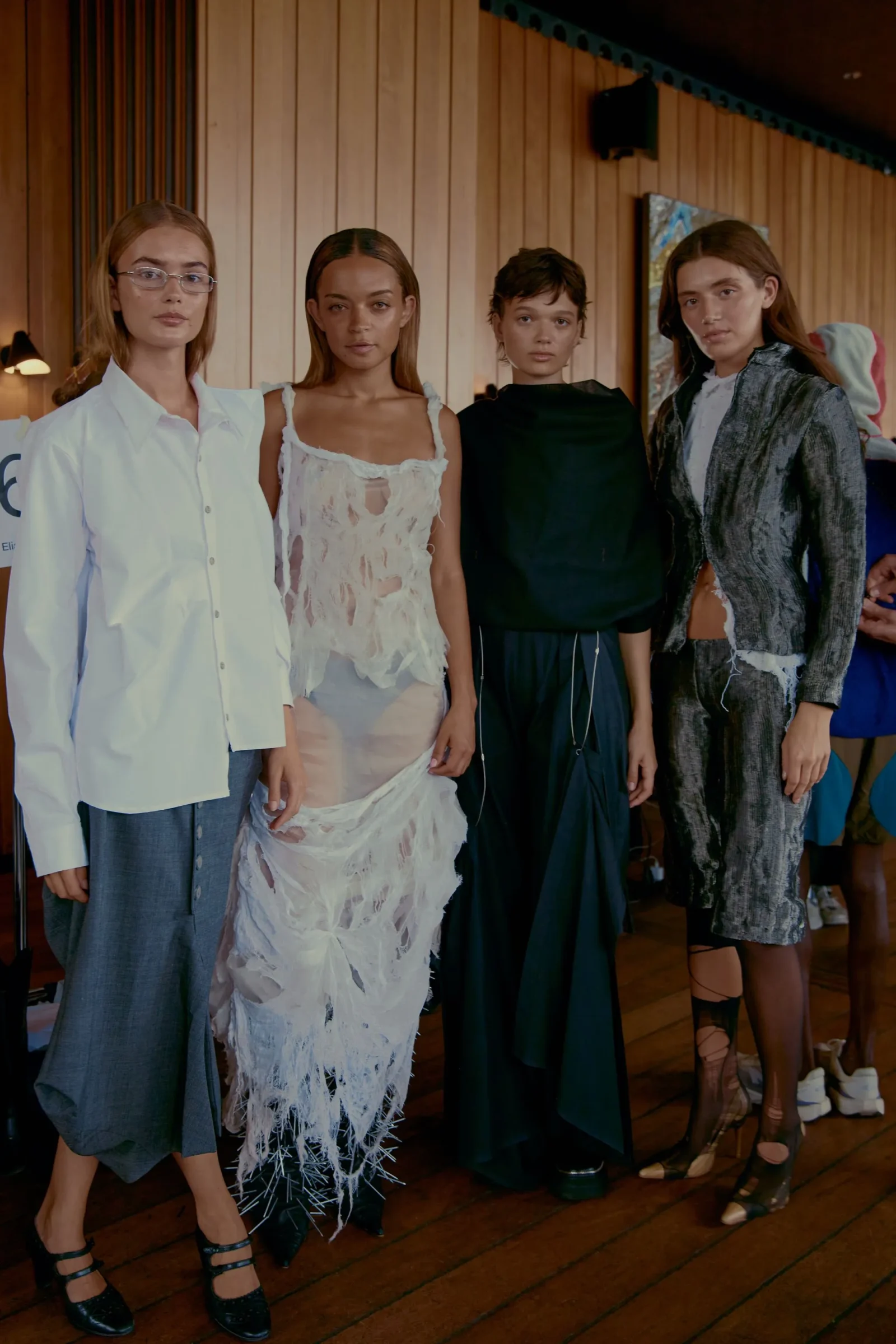 Four models in designer outfits pose together indoors in a wood-paneled room, backstage at a fashion event.