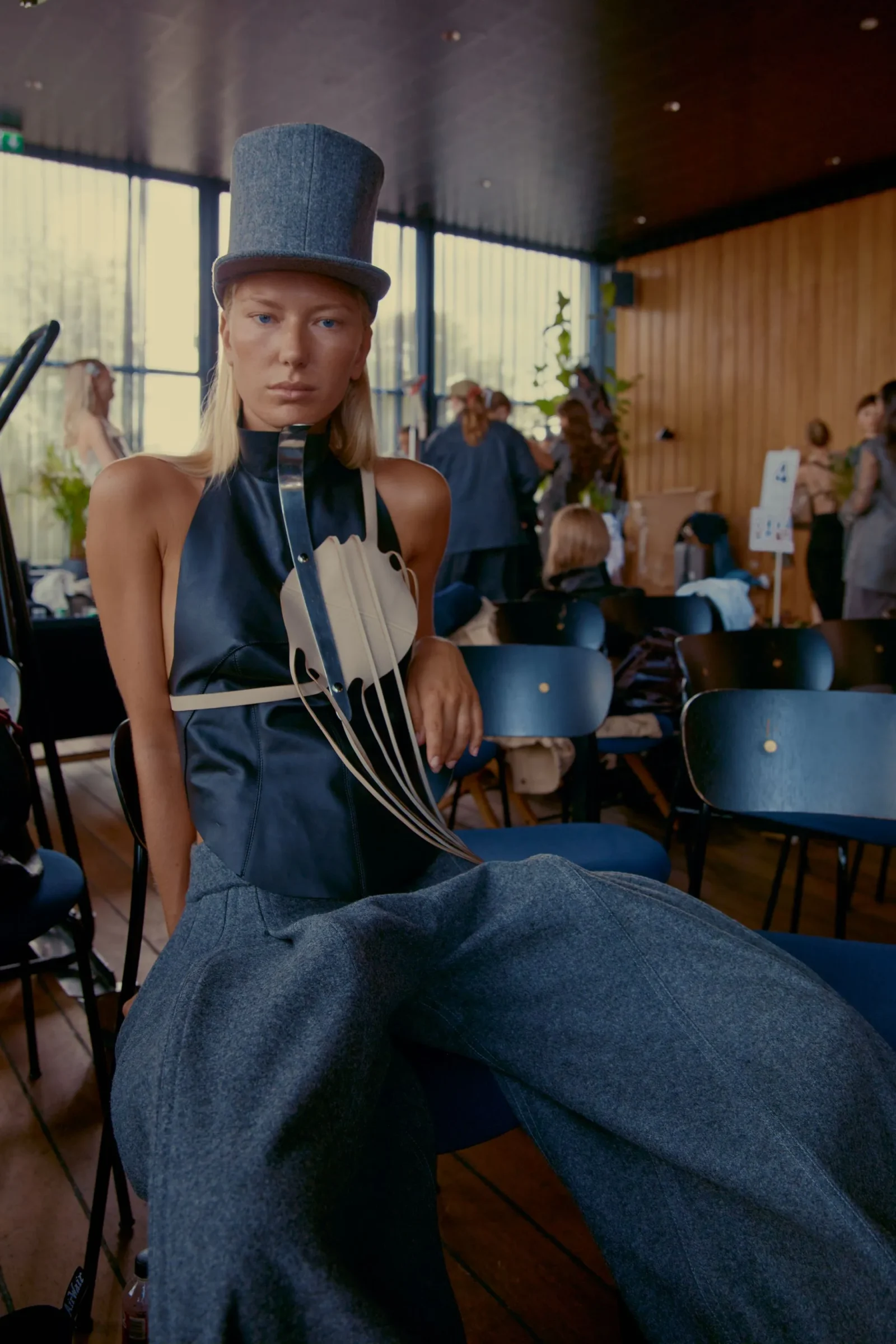 Female model wearing a tall blue top hat and a navy sculptural outfit in a busy indoor event space with wooden walls and blue chairs nearby.