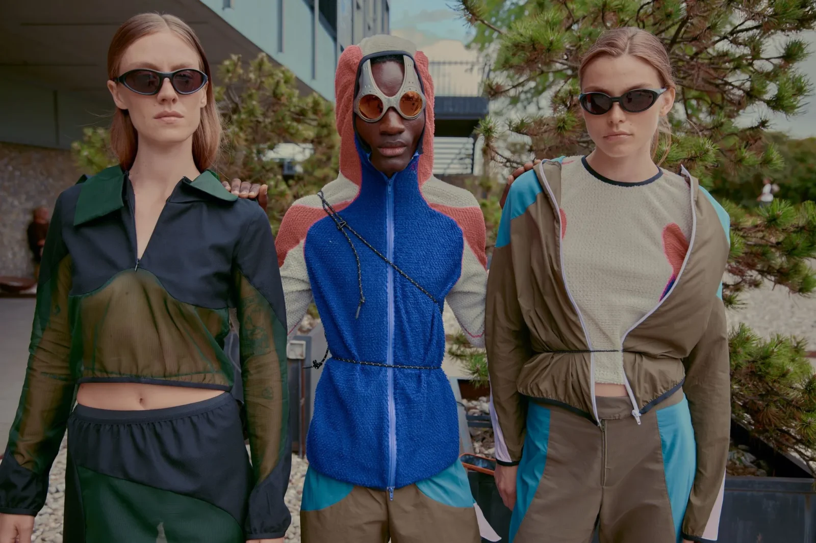 Three models stand outdoors wearing bold, color-block streetwear and dark sunglasses.