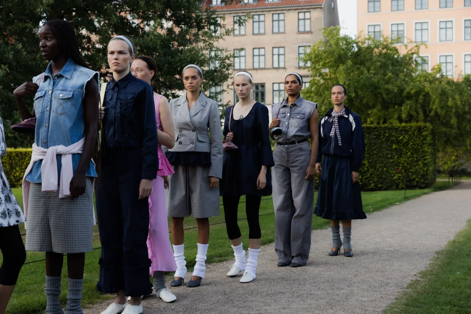 Group of diverse women modeling outfits outdoors along a park path with apartment buildings in the background, wearing varied fashion styles.
