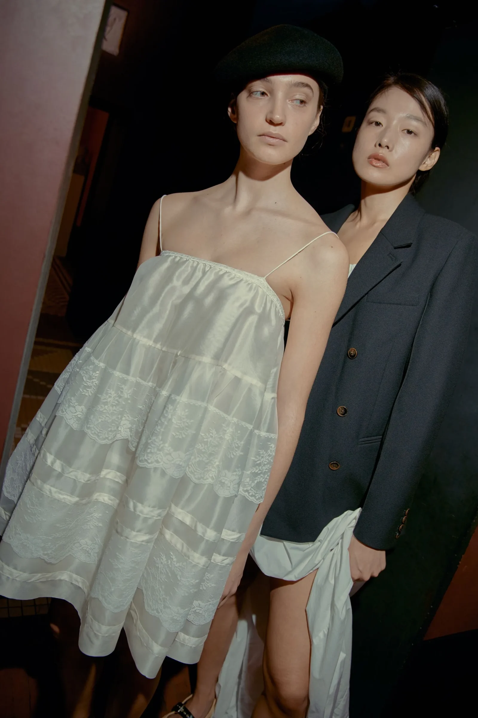 Two models backstage wearing chic outfits: one in a white lace dress with a black beret, the other in a dark blazer.