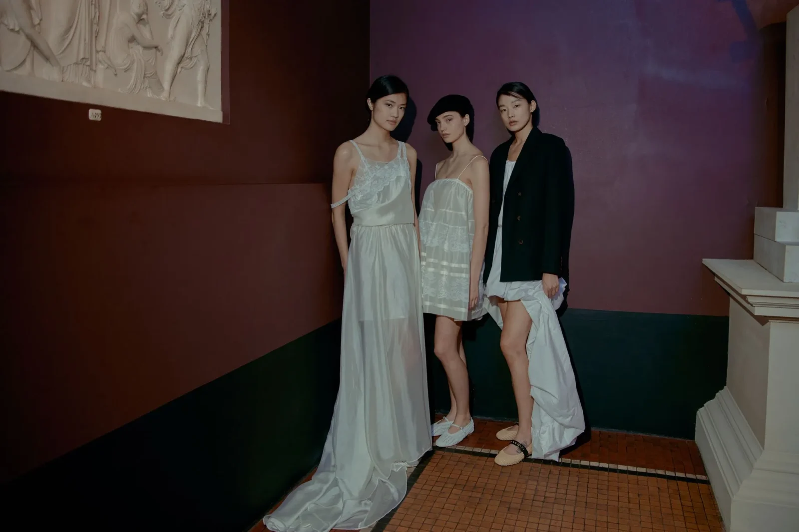 Three models in pale gowns pose together against a burgundy wall in a gallery-like hallway with a marble relief on the left.