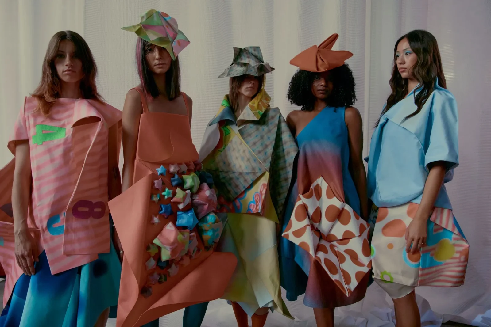 Five models stand side by side in bold, multicolored avant-garde outfits and sculptural hats during a fashion photoshoot in a pale setting.