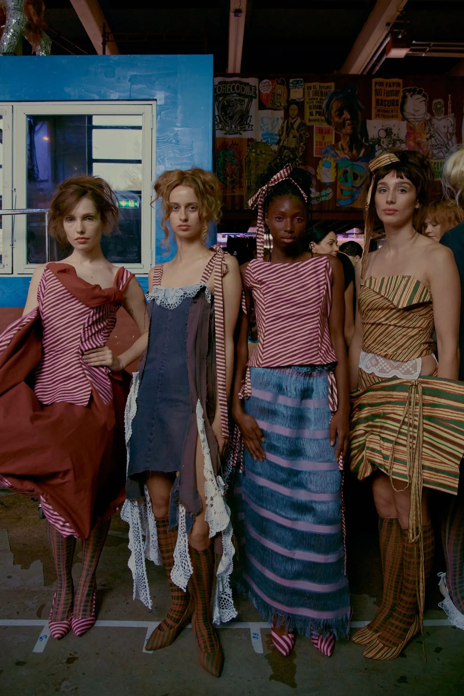 Four female models stand together in striped and lace-trimmed dresses, backstage at a fashion show with a colorful mural behind them.