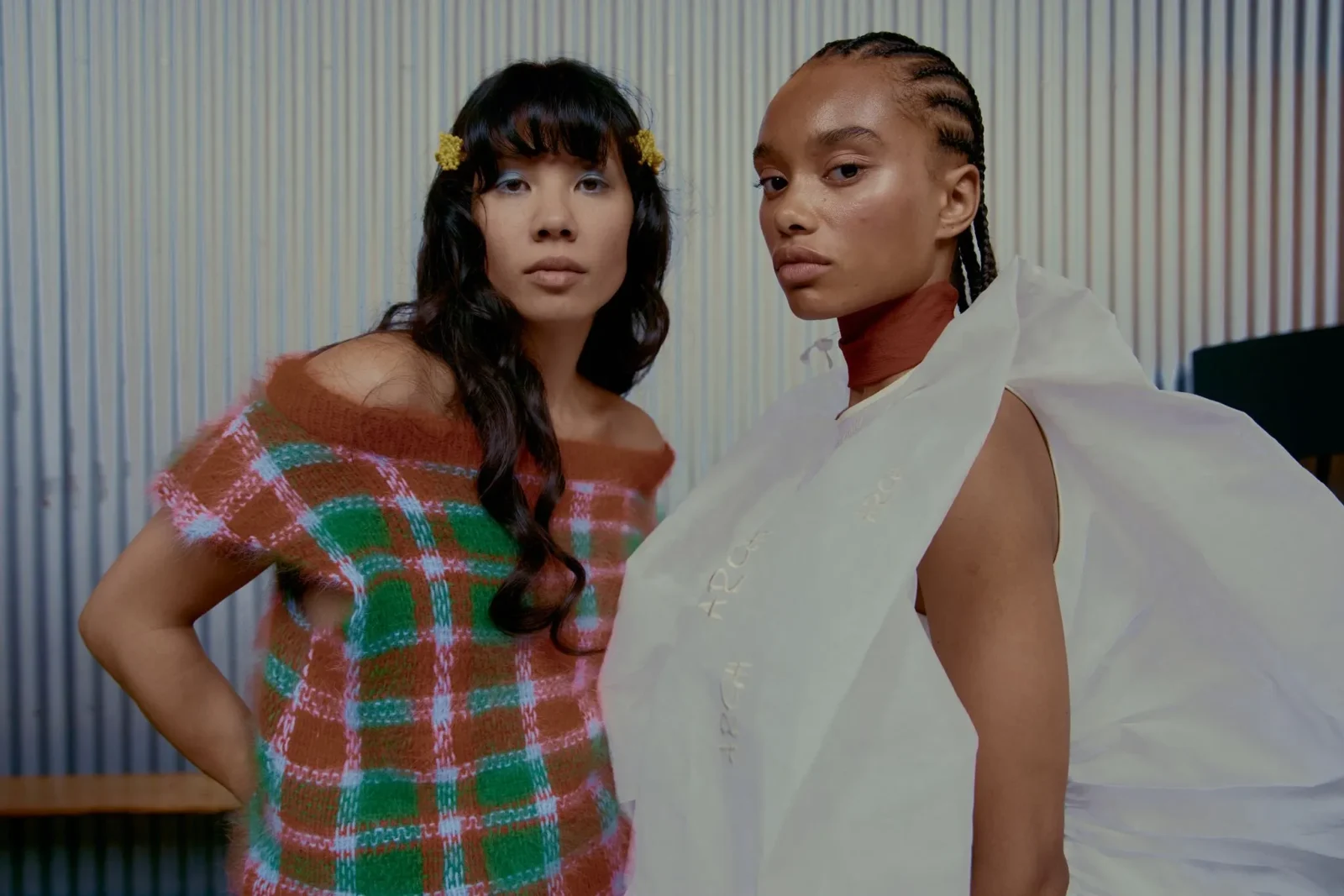 Two women pose together in a studio: left in an orange-green plaid sweater, right in a white, draped garment with braided hair, striped backdrop behind them.