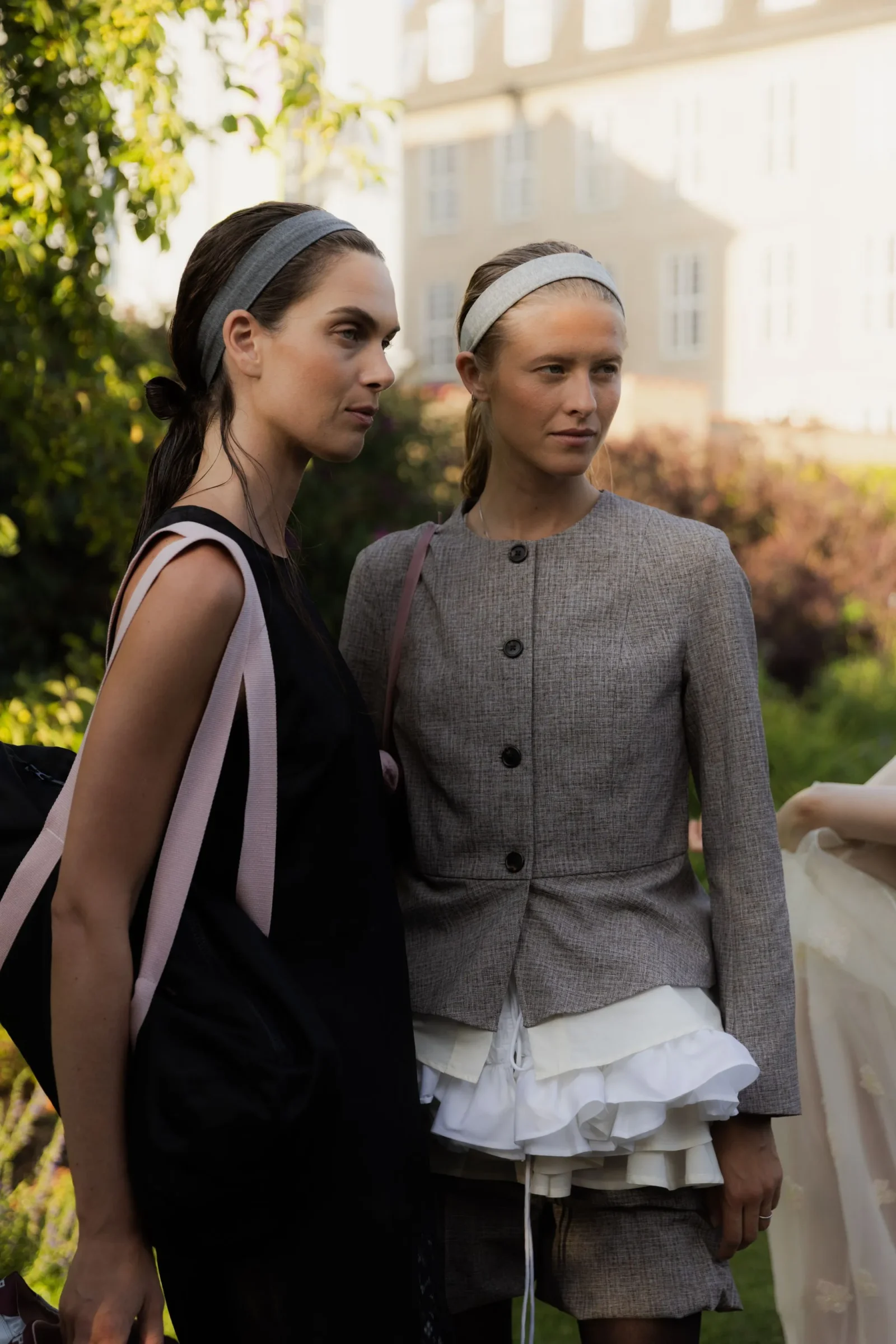 Two women stand outdoors in a garden setting, wearing stylish outfits and gray headbands, looking off to the side.