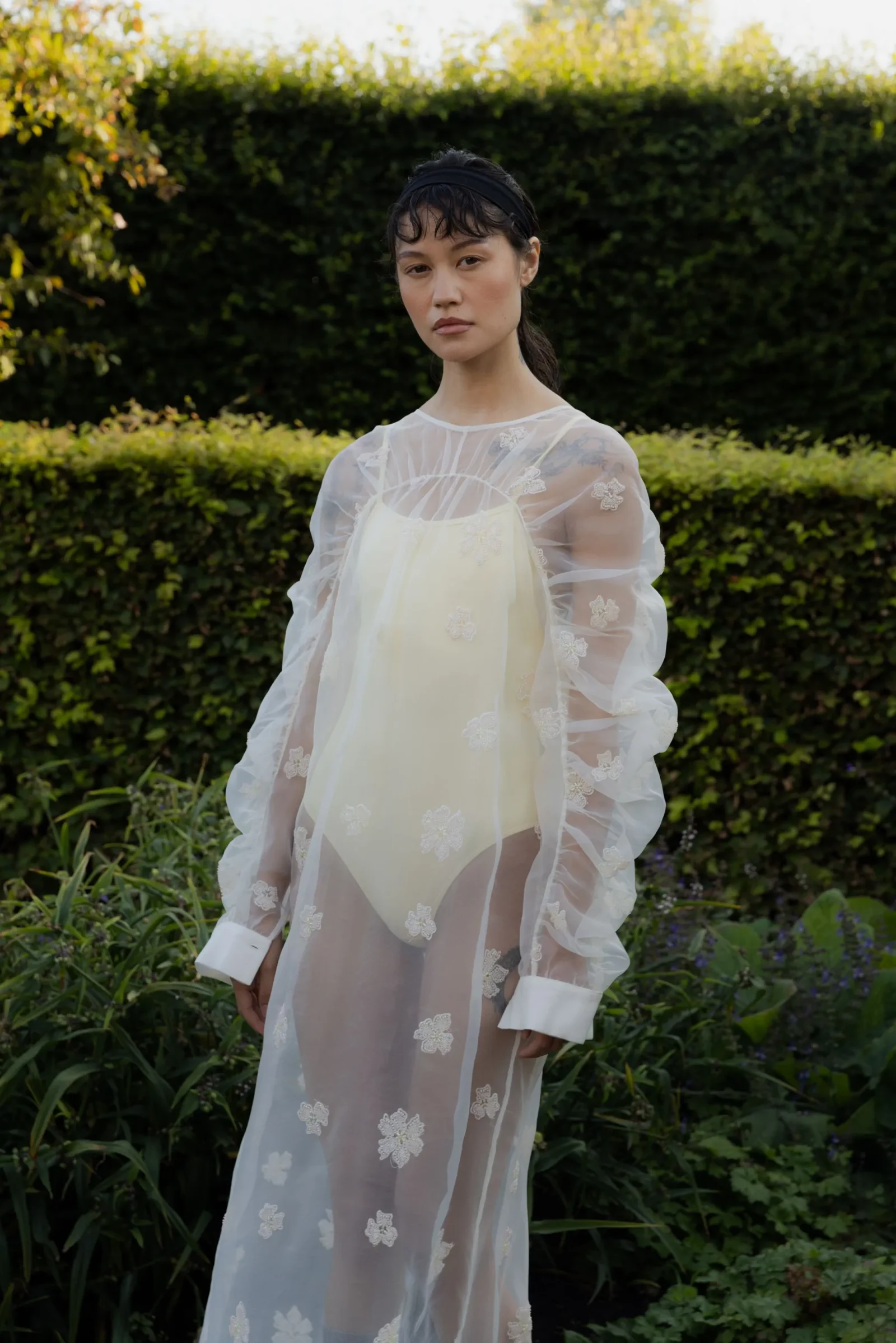 Woman standing in a garden wearing a sheer white floral lace dress, looking at the camera with a neutral expression.