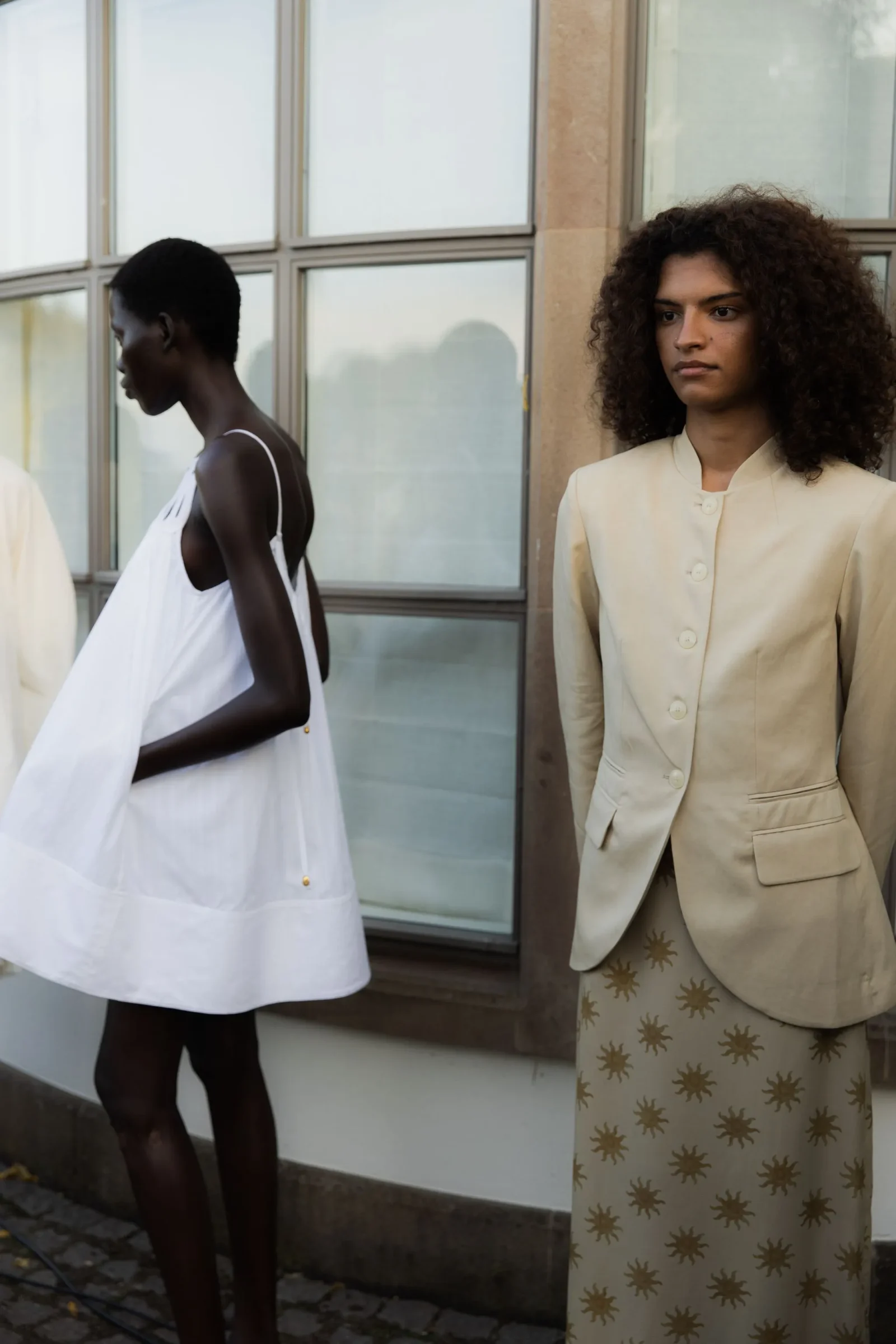 Two models pose outdoors beside a glass wall; one in a white flowing dress, the other in a cream blazer with a star-patterned skirt.