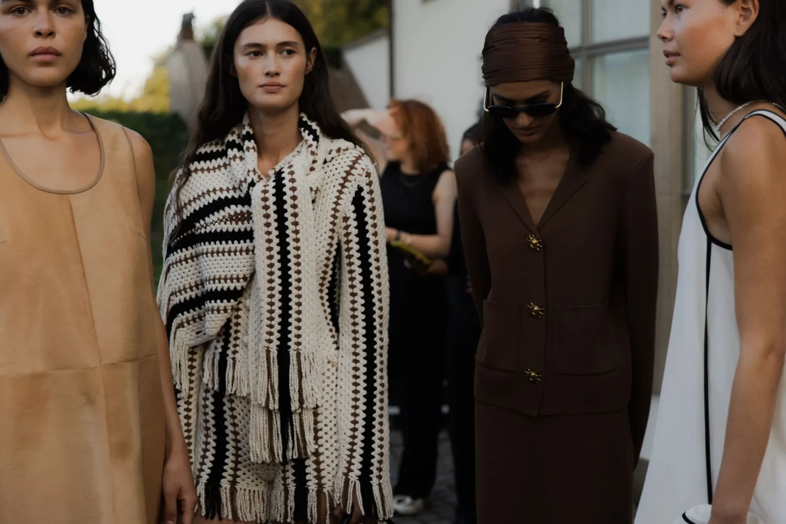 Group of fashion models standing outdoors in stylish neutral-toned outfits: beige sleeveless dress, crochet-striped cardigan, brown coat, and white dress with black trim.