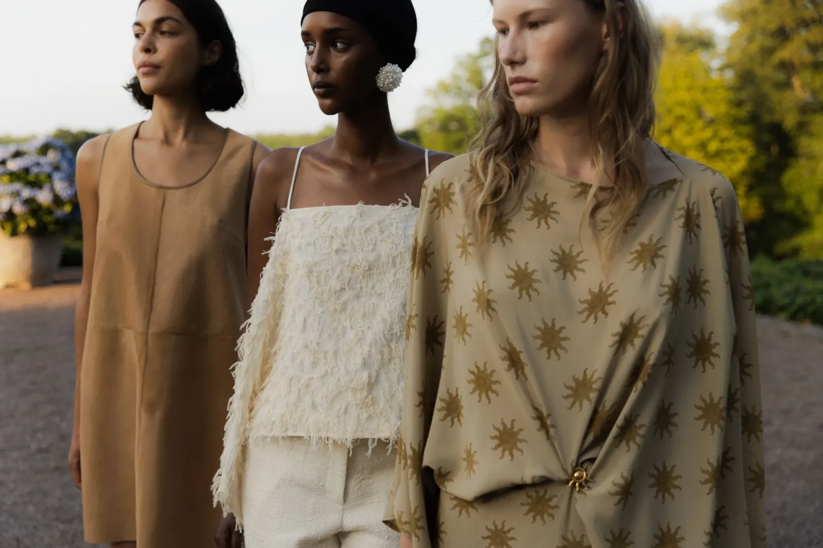 Three women standing side by side outdoors in warm evening light; left in a tan sleeveless dress, center in a white textured dress with a headwrap, right in a beige patterned dress with sun motifs.