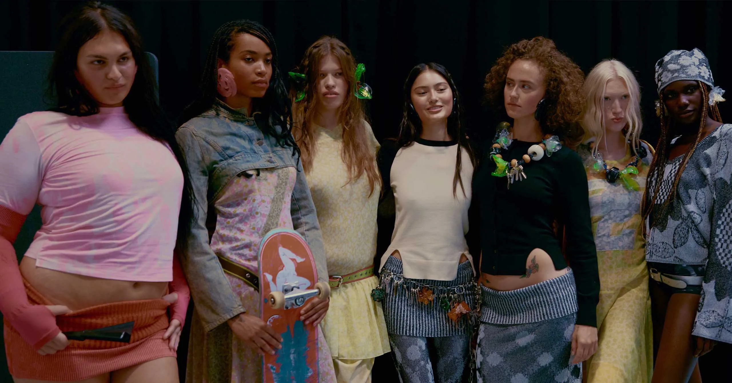 Group of diverse young people posing on a stage in colorful, eclectic outfits and accessories against a dark backdrop.