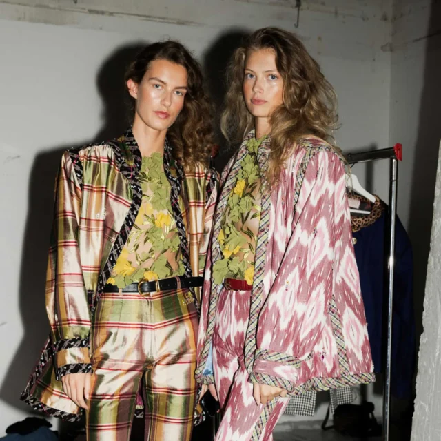 Two models stand side by side in bold patterned outfits, backstage setting with a gray wall behind them.
