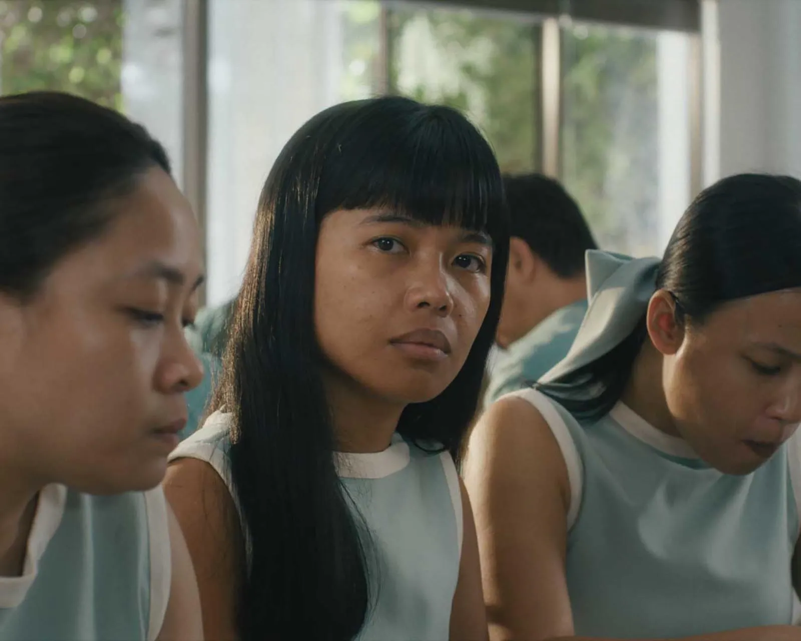 Three women in light blue uniforms sit closely together; the center woman looks toward the camera with a neutral expression.