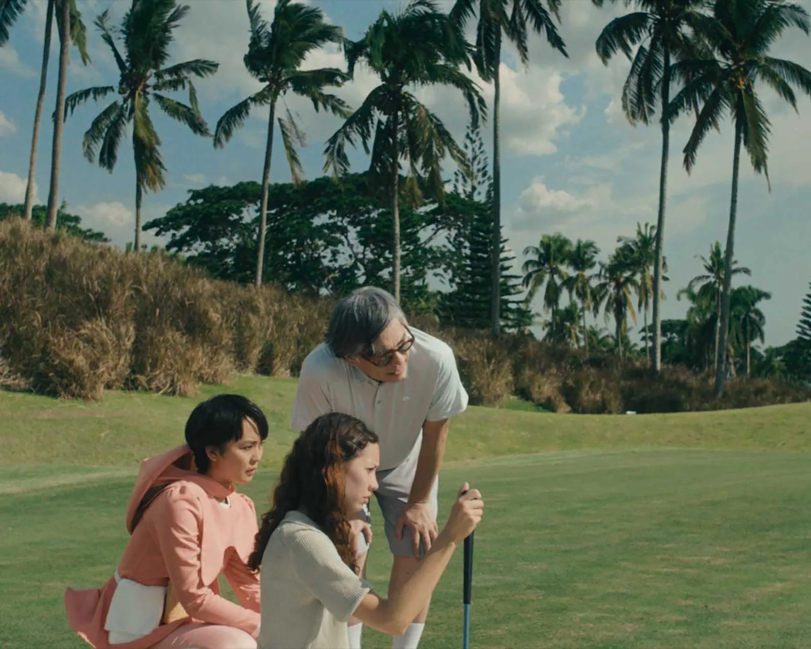 Group of people outdoors on a sunny, grassy field with palm trees in the background; an older man stands beside two seated individuals, guiding them with a golf club or stick.
