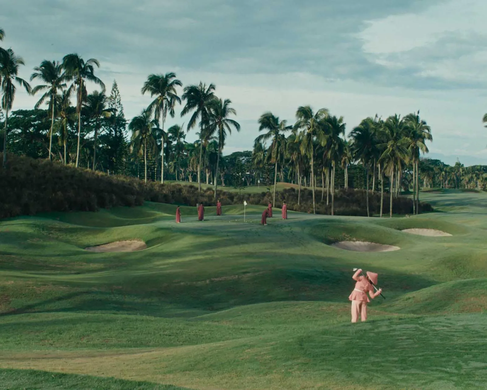 Golfers in pink outfits on a green golf course with tall palm trees in the background.