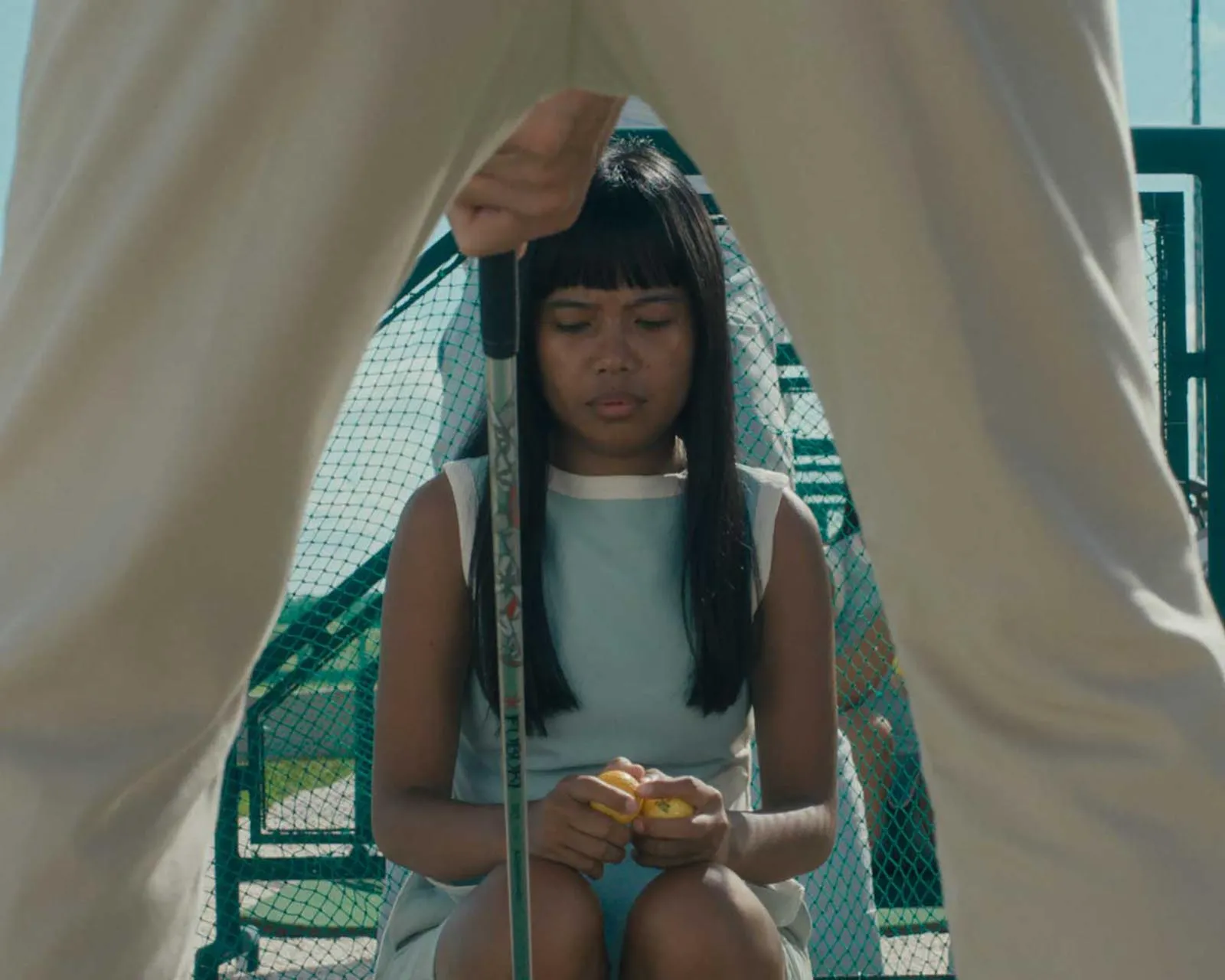 Girl with long dark hair sits cross‑legged on a playground, peeling a yellow fruit while an adult in beige pants stands over her.