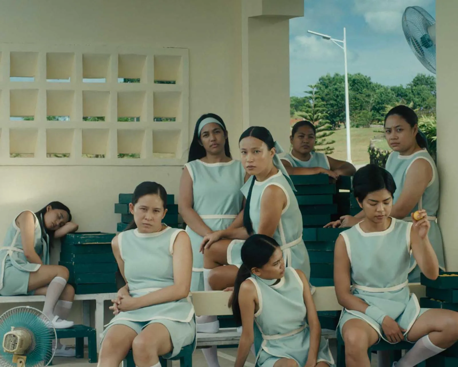 Group of women in matching pale blue uniforms sit and stand around blue bleachers under a pavilion, looking toward the camera/around the area.