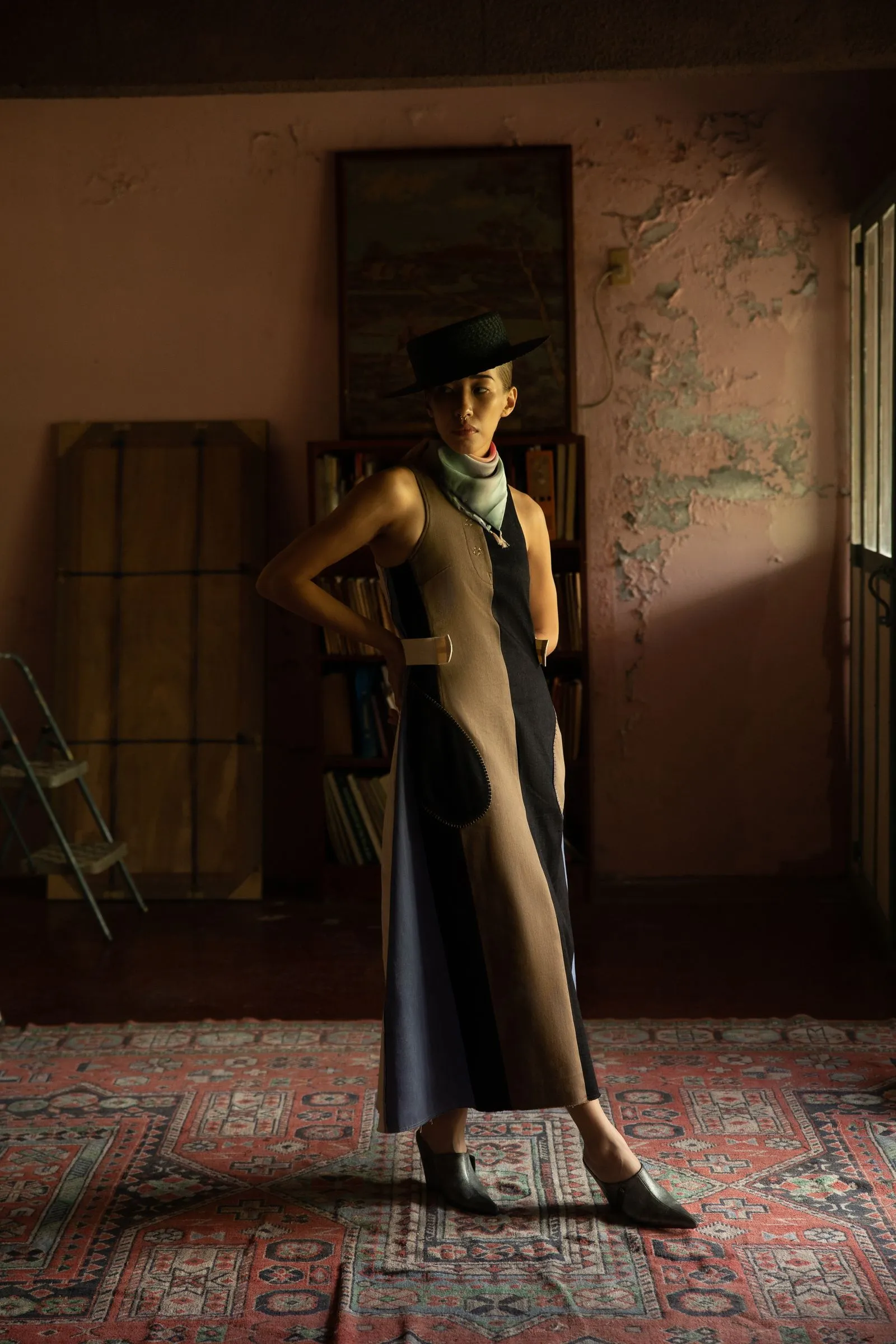 Woman posing in a color-block sleeveless dress and wide-brim hat, standing in a dim, pink-walled room with a patterned rug.