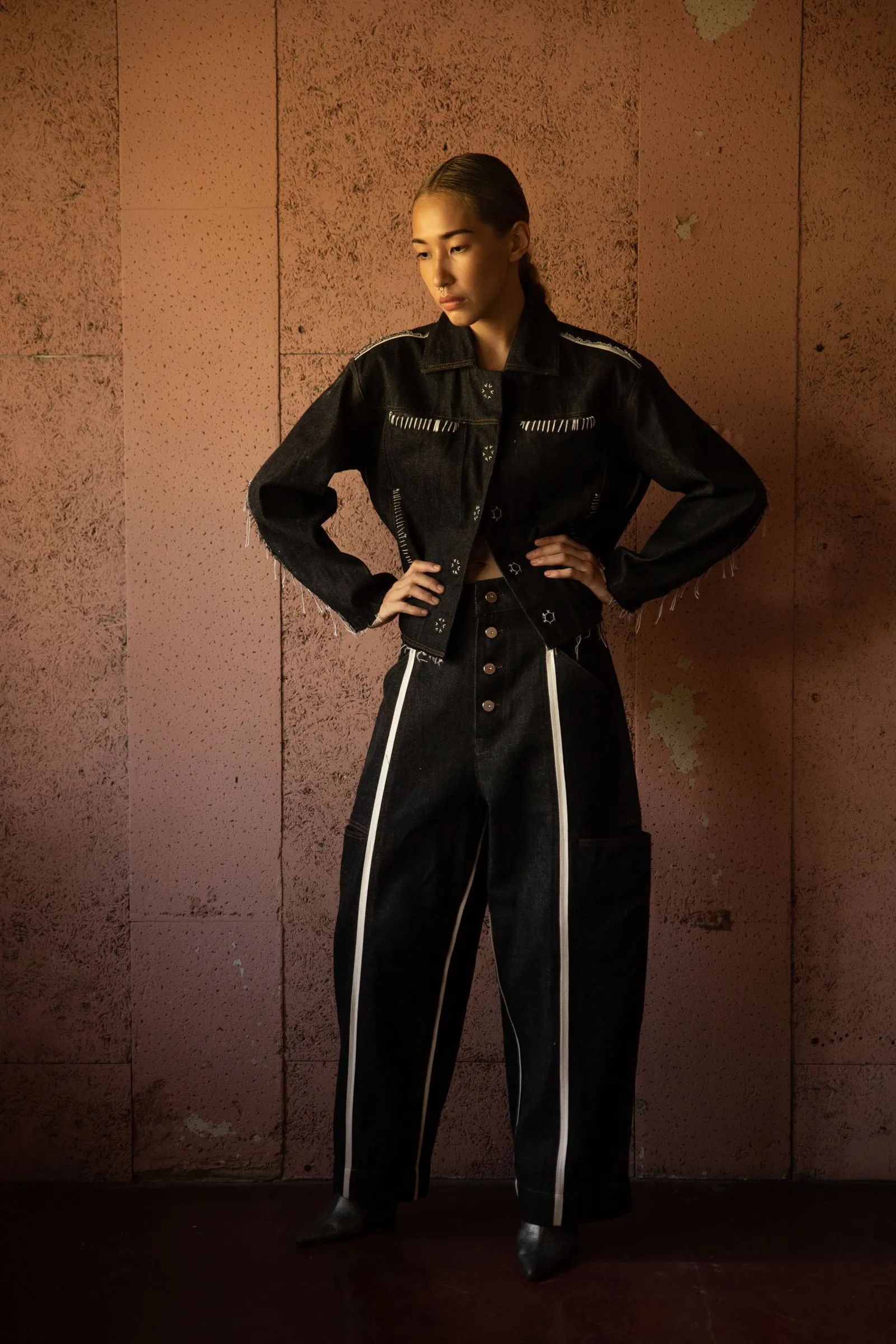 Woman in a black denim outfit with white stitching stands with hands on hips against a pink textured wall.
