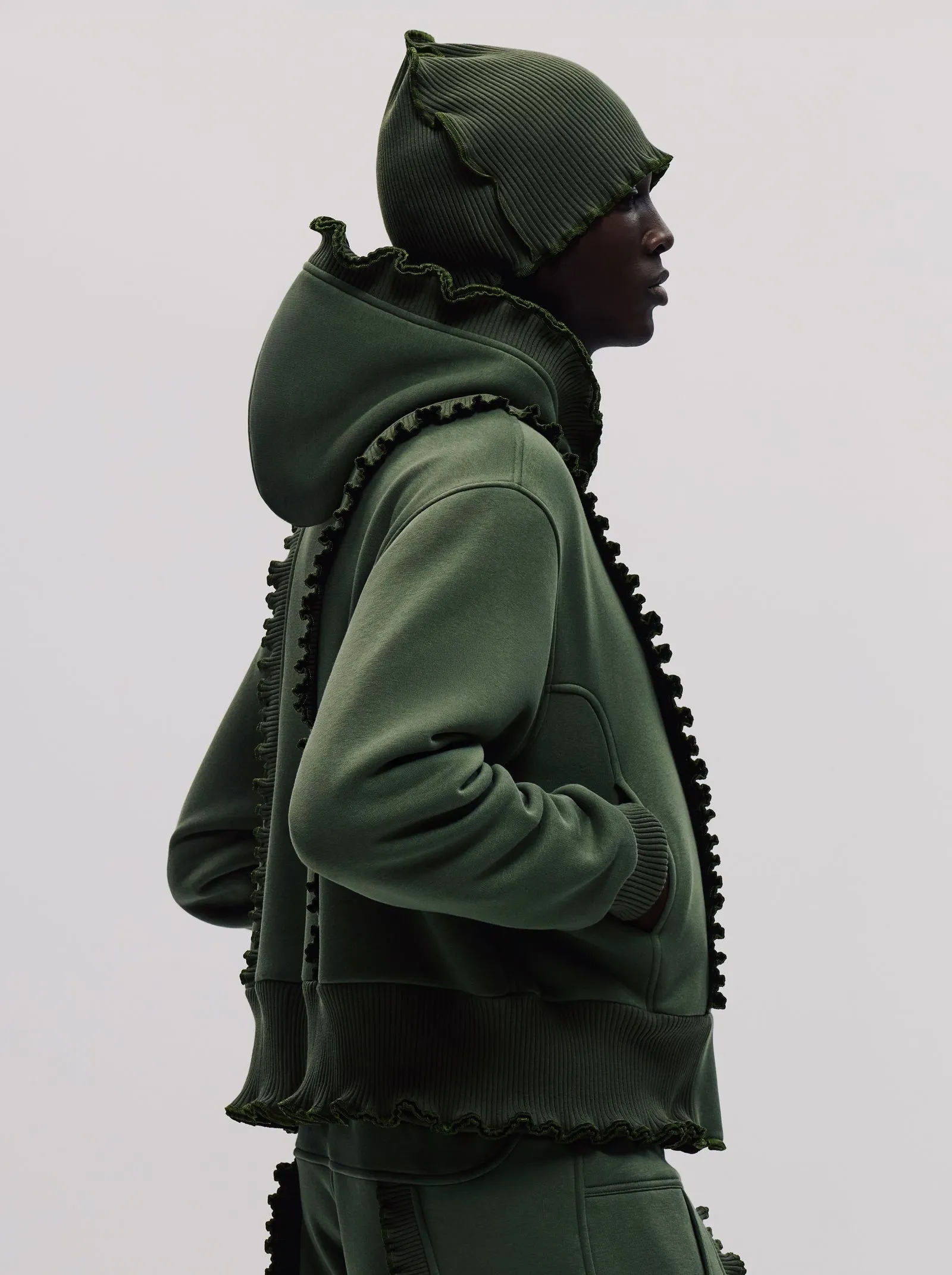 Side profile of a person wearing an oversized olive-green hoodie with a ruffled edge along the hood and seams.