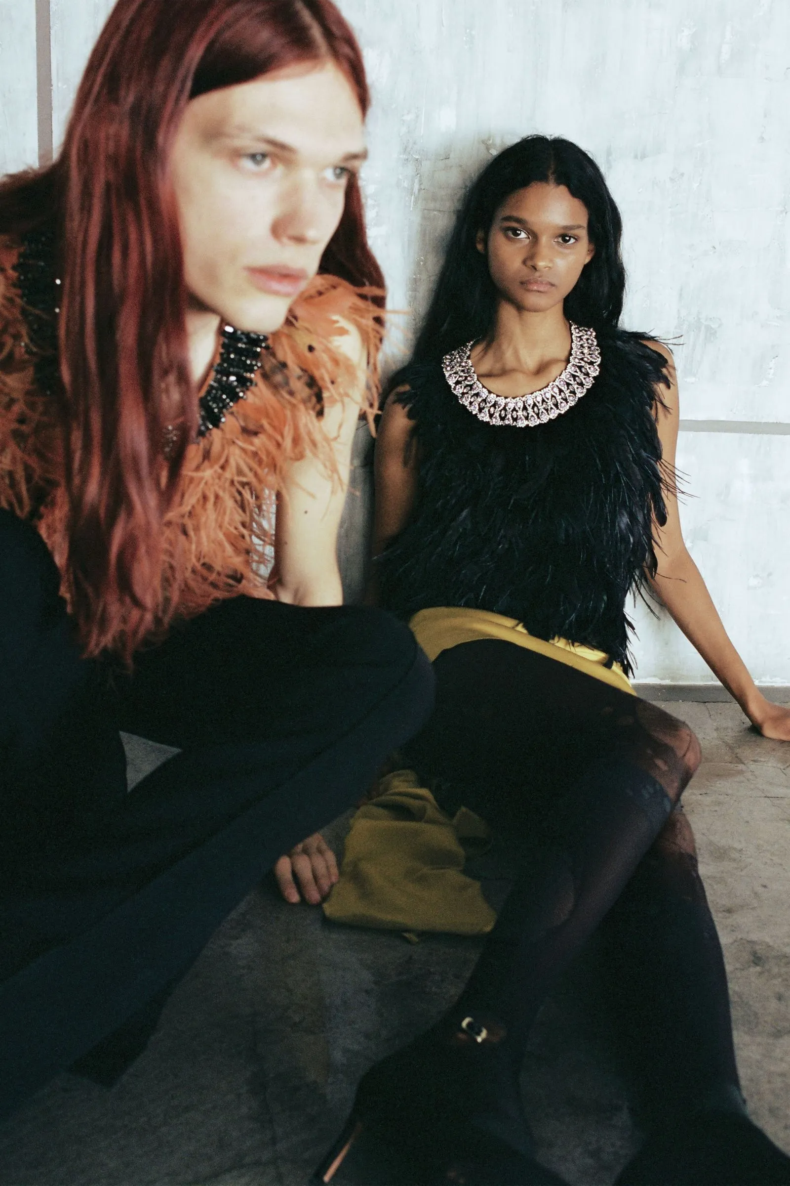 Two models sit on the floor against a gray wall; one with red hair and a feathered brown top looks away, the other with black hair in a jeweled feathered dress faces the camera.