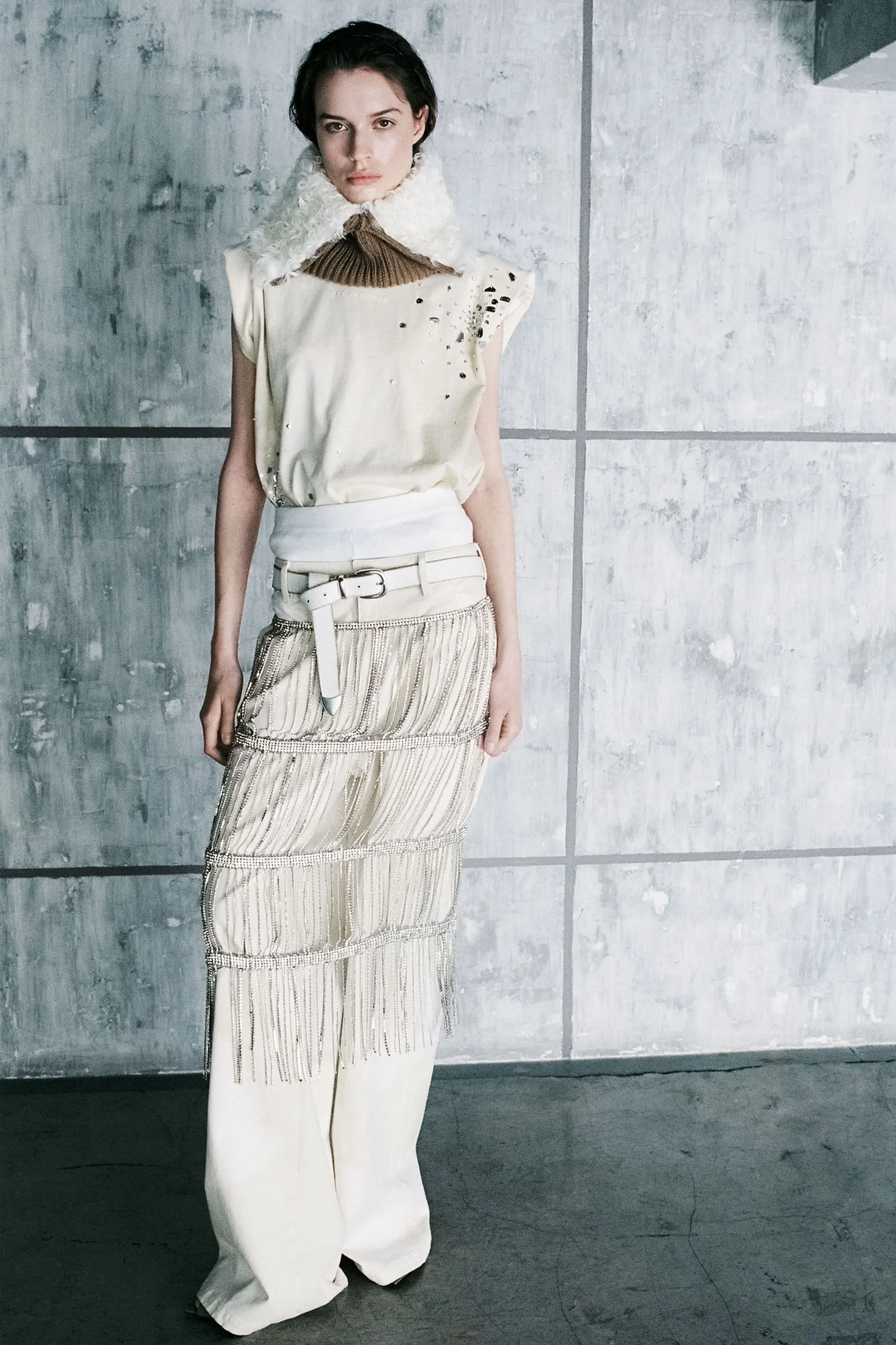 Fashion model wearing an ivory sleeveless top, fringed white skirt, and a fluffy scarf, posed against a gray concrete backdrop.
