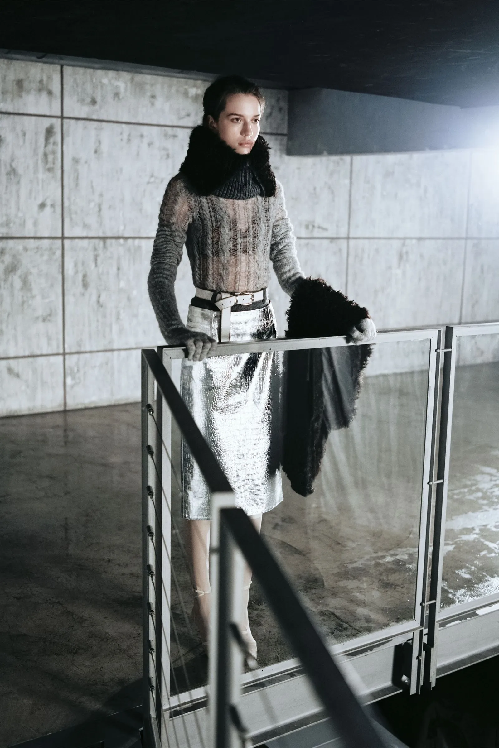 Woman in a metallic silver skirt and chunky knit sweater stands at a glass railing in an industrial concrete interior, gazing to the side.